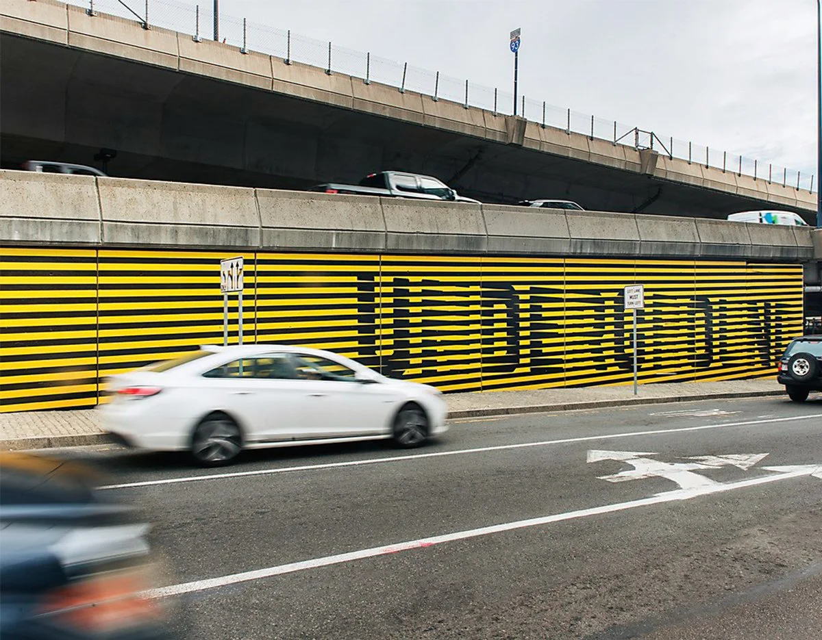 Cars driving past a black-and-yellow striped retaining wall beneath an Interstate 93 overpass in Boston.