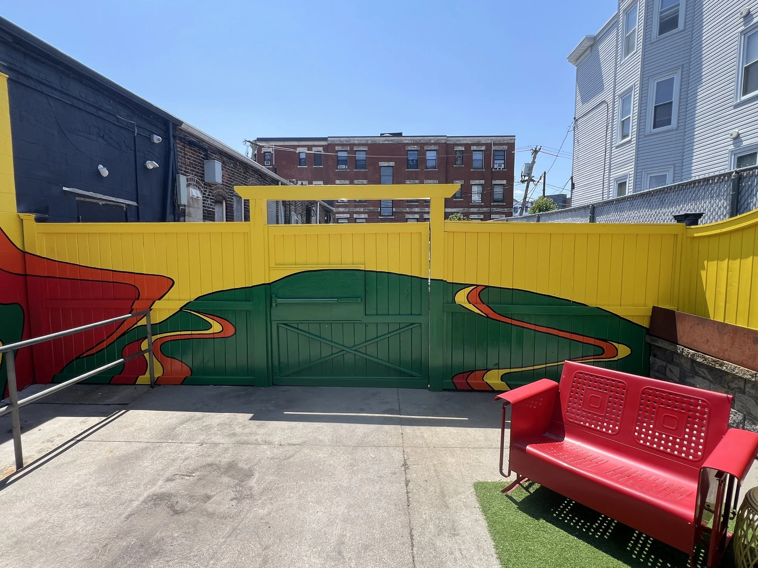 Colorful abstract mural on a courtyard wall between urban buildings, with red outdoor seating in the foreground.