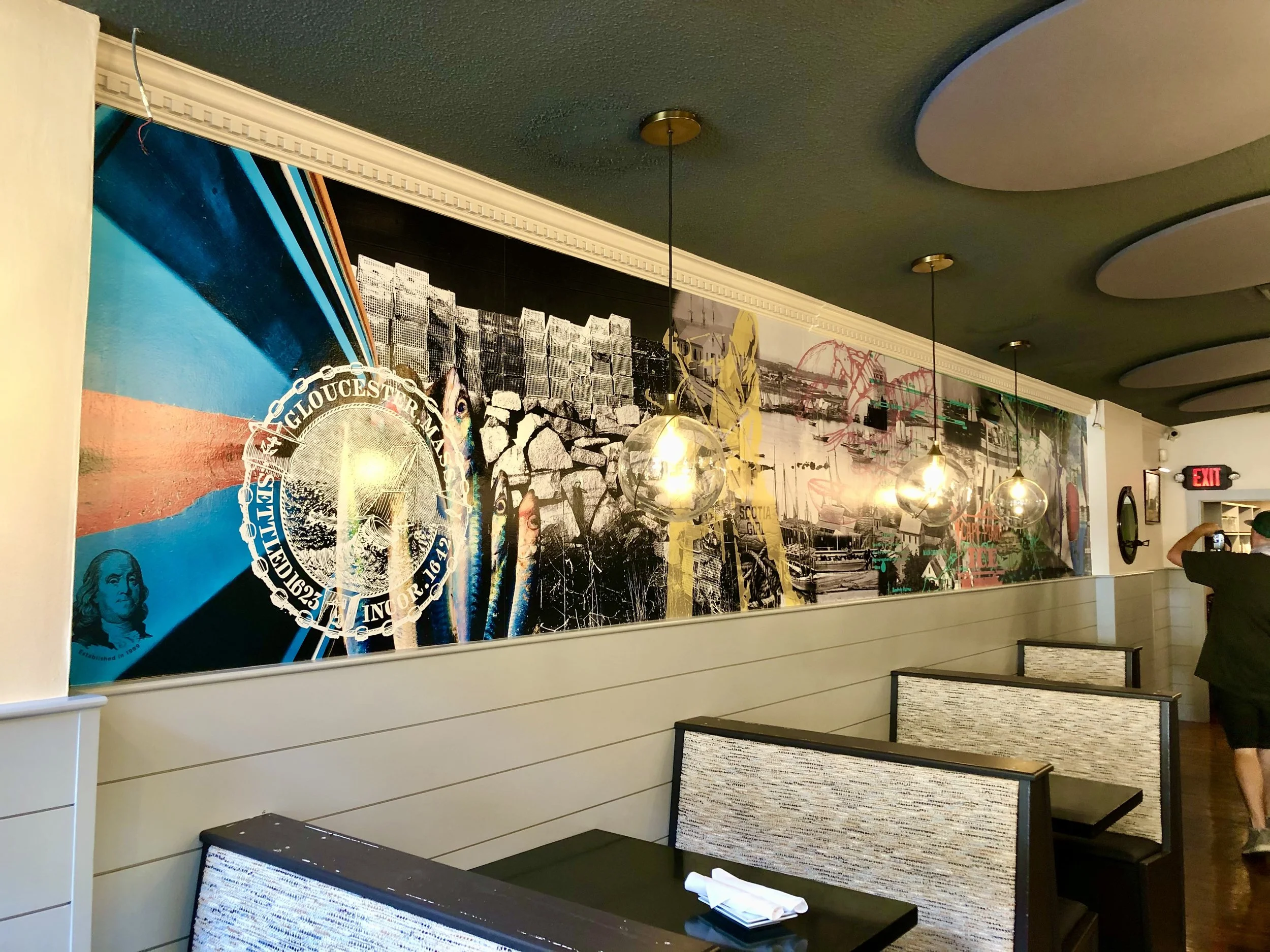 Restaurant dining room with nautical murals, booth seating, globe pendant lights, and an illuminated exit sign.