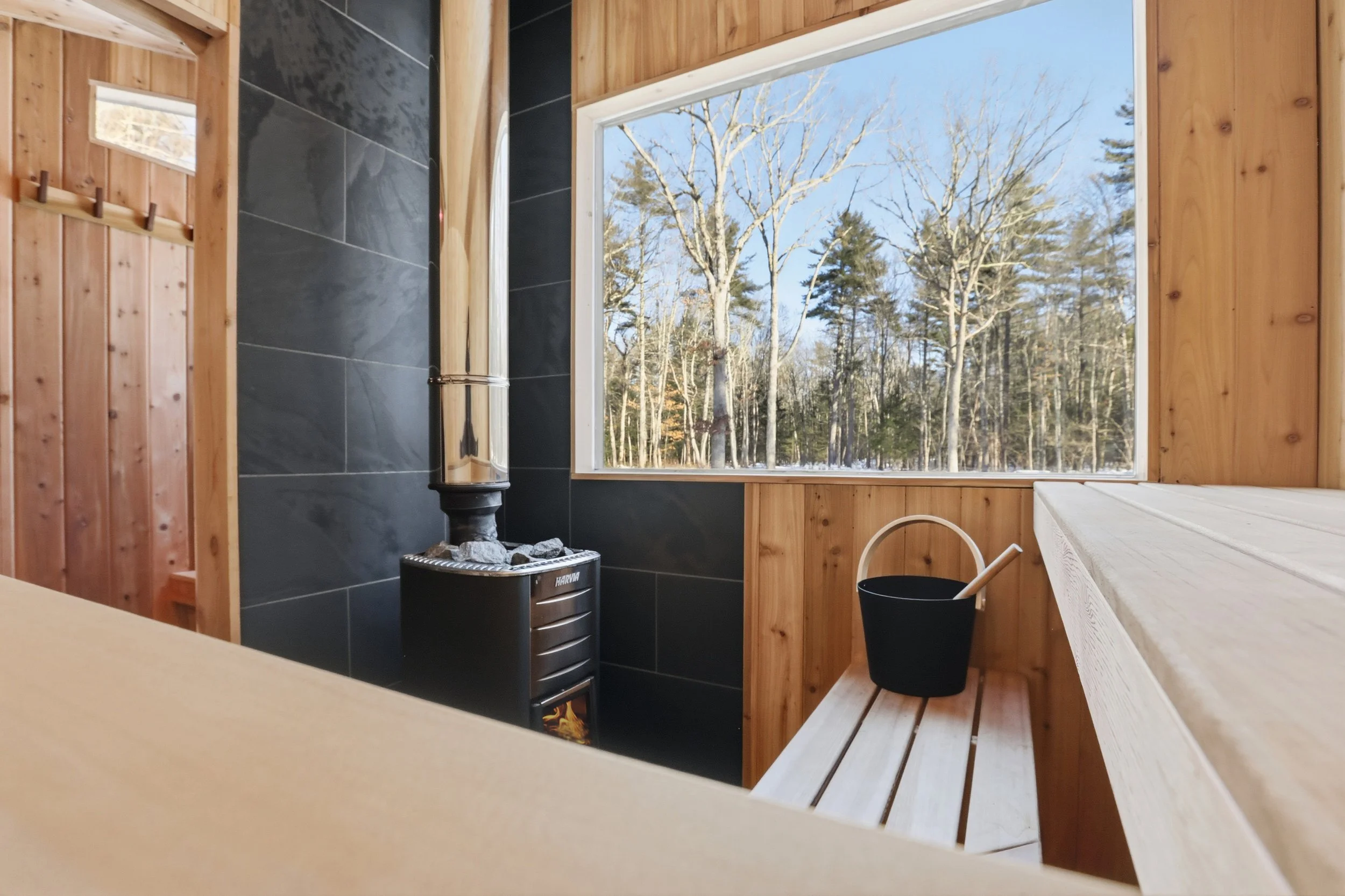 Wood-paneled sauna with a Harvia wood-burning stove, benches, bucket and ladle, and a large window overlooking a snowy forest.