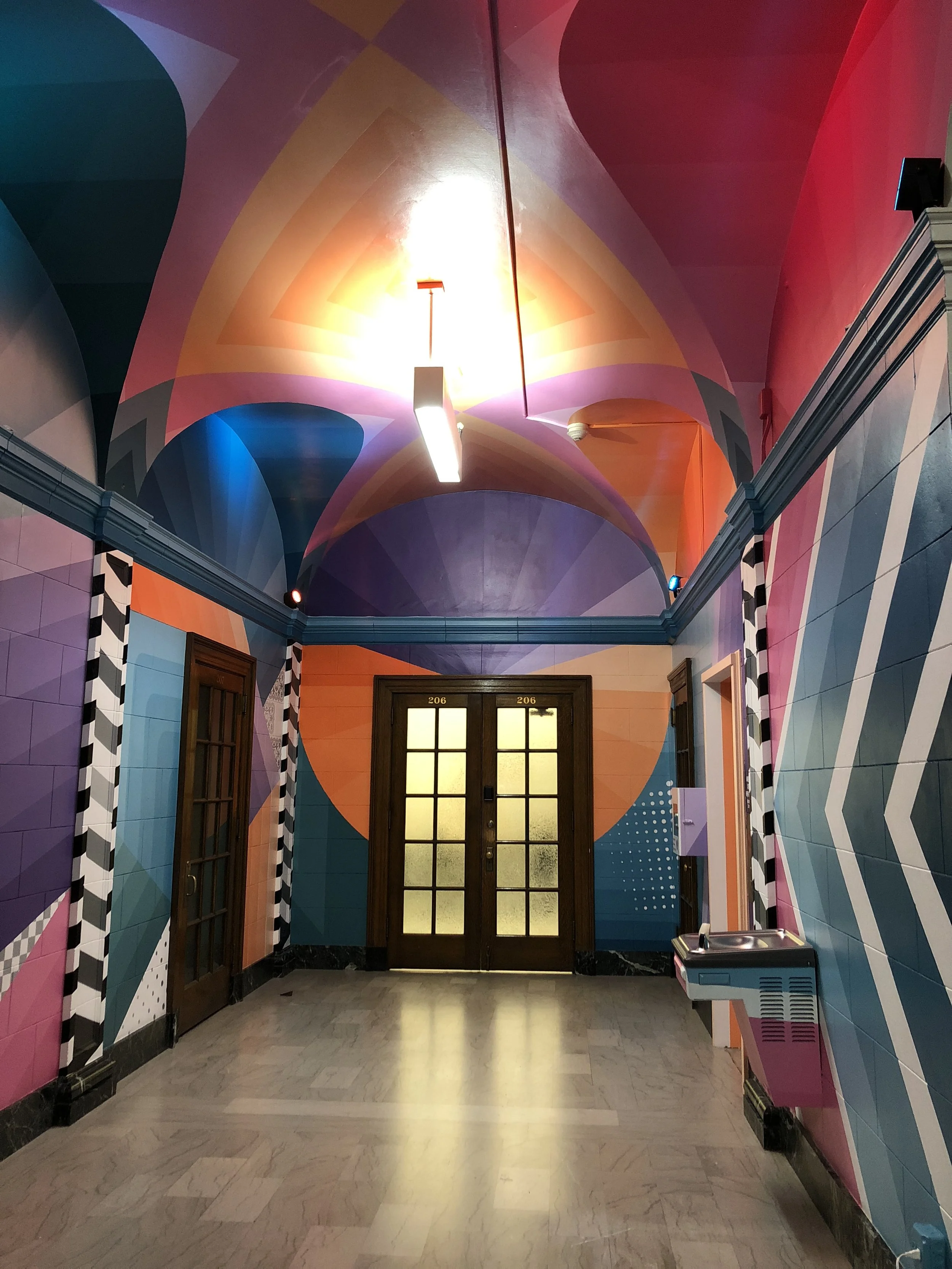 Colorful geometric mural in an interior hallway with arched ceiling, pendant lights, double doors labeled 206, and a drinking fountain.