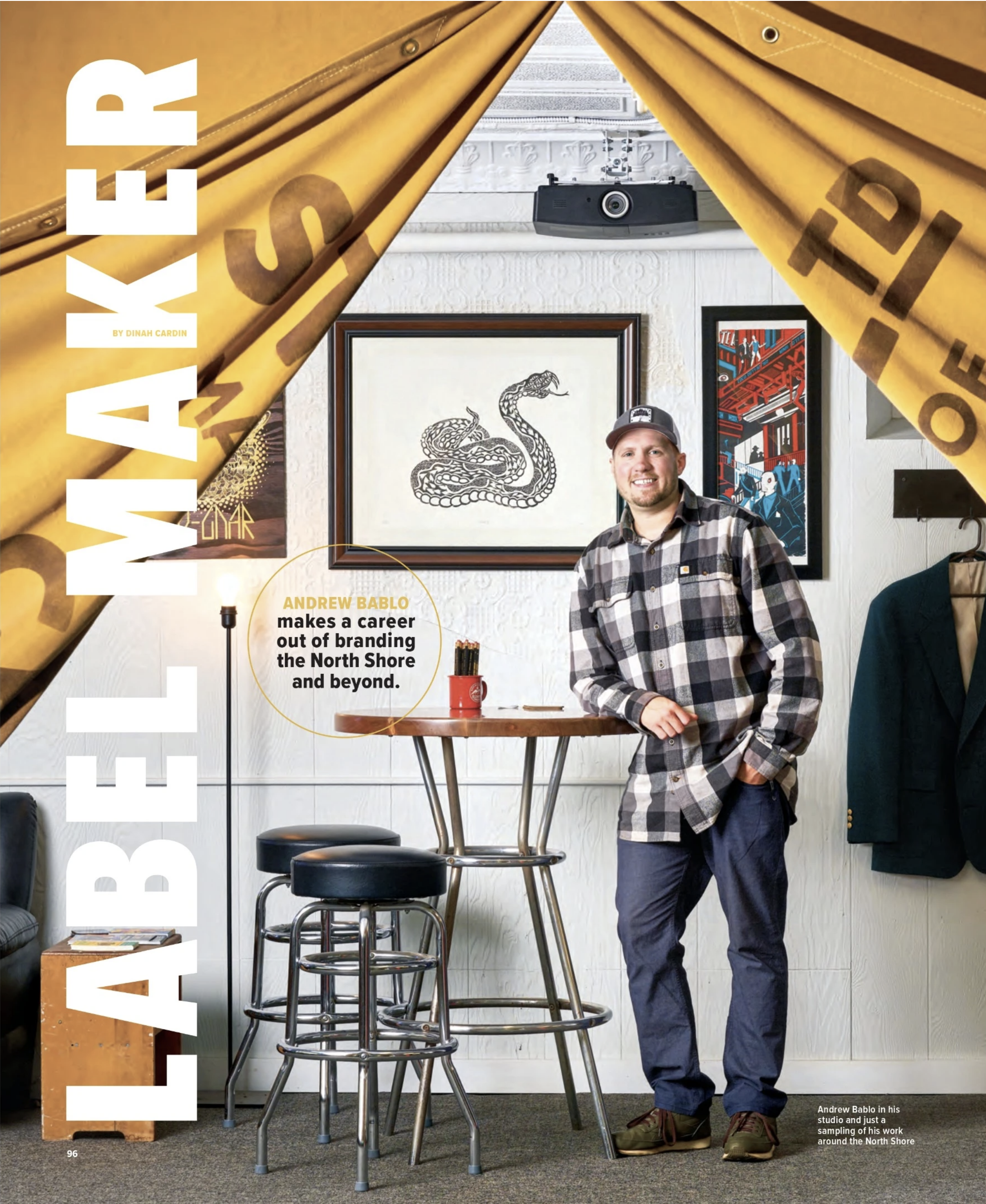 Magazine feature portrait of branding designer Andrew Bablo standing in his studio beside bar stools, artwork, and draped yellow banners.