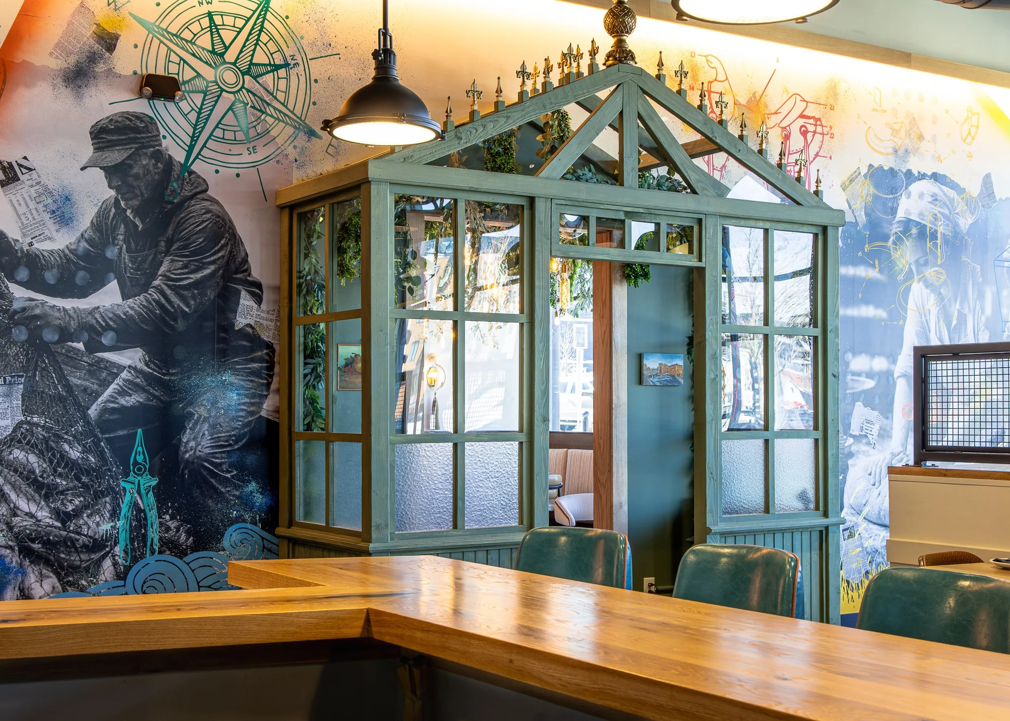 Eclectic restaurant interior with green glass-paneled divider, wooden tables, leather chairs, nautical murals, and industrial pendant lights