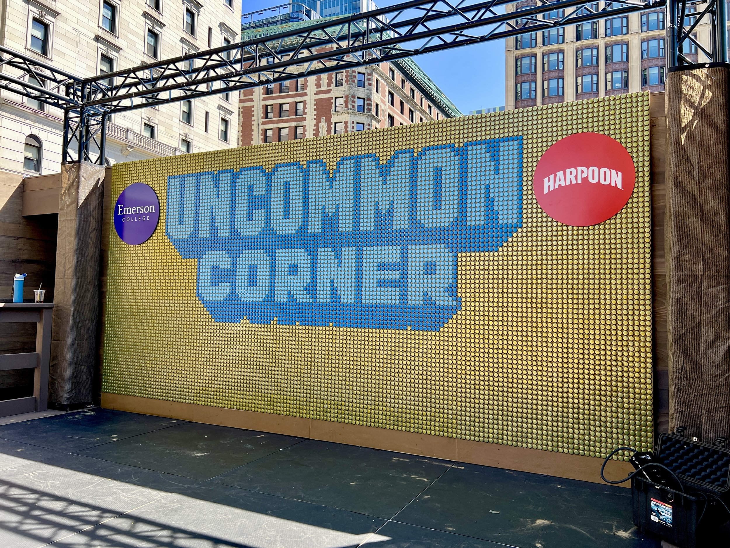 Harpoon beer promotional mural made of colored bottle caps, with Emerson College and Harpoon logos, surrounded by Boston buildings.
