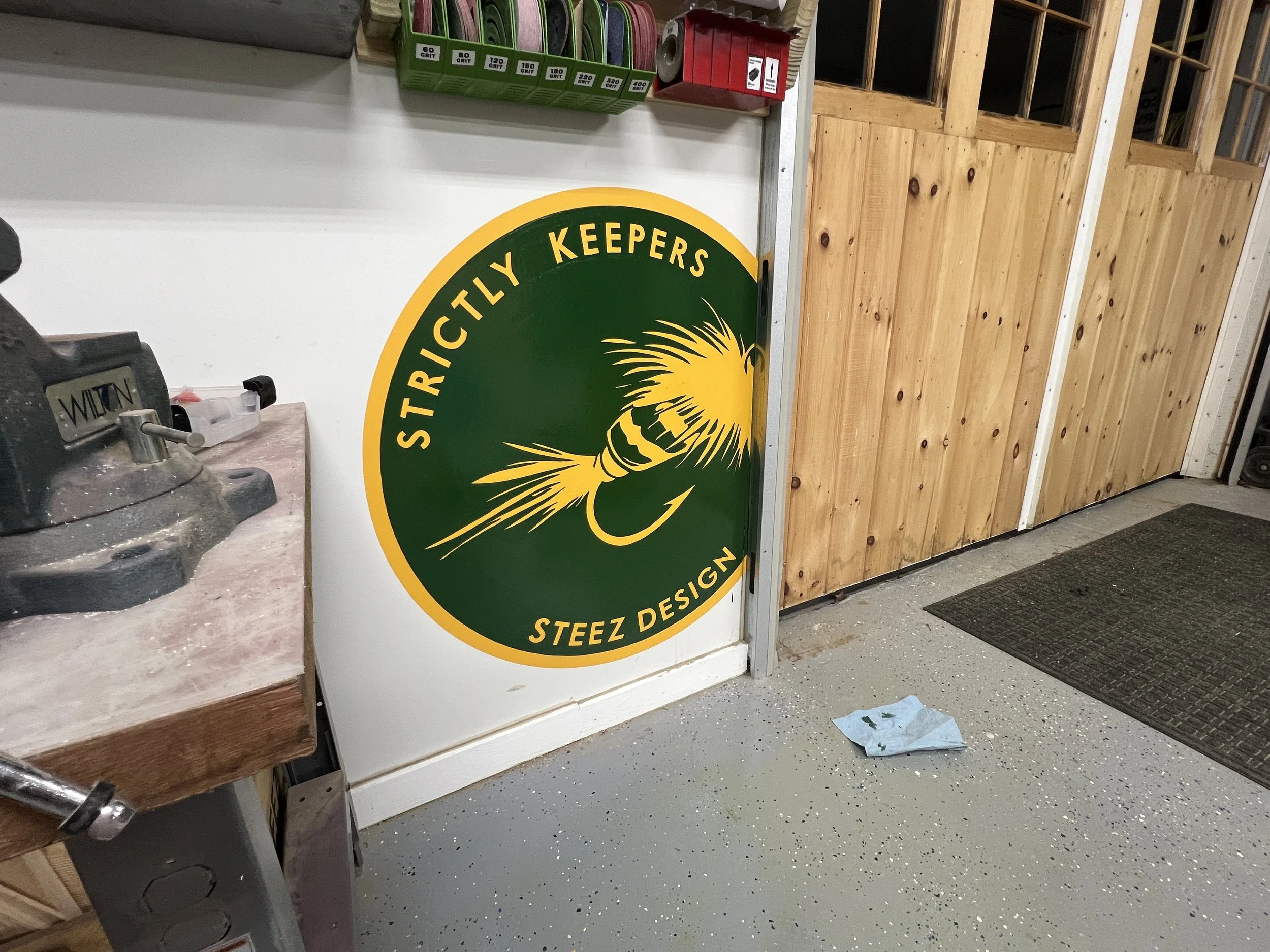 Green and yellow Strictly Keepers Steez Design sign displayed in a workshop with sanding belts and woodworking equipment