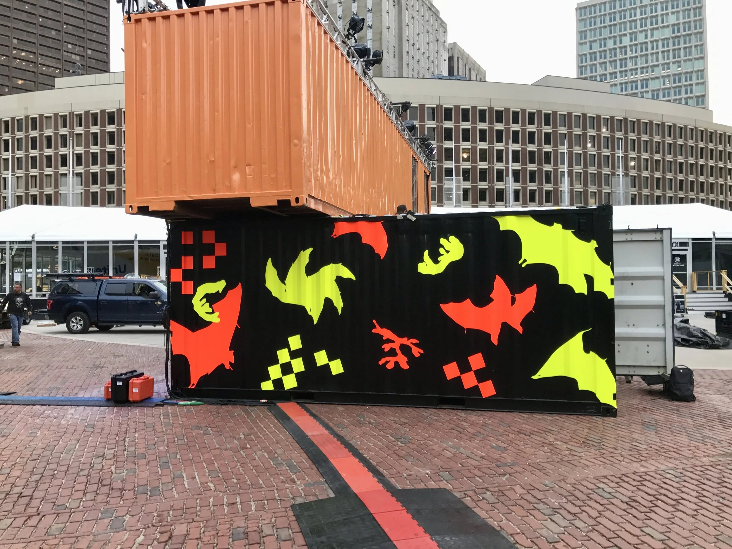 Colorful outdoor event installation made from shipping containers in a downtown plaza, with neon animal silhouettes and city buildings in the background.
