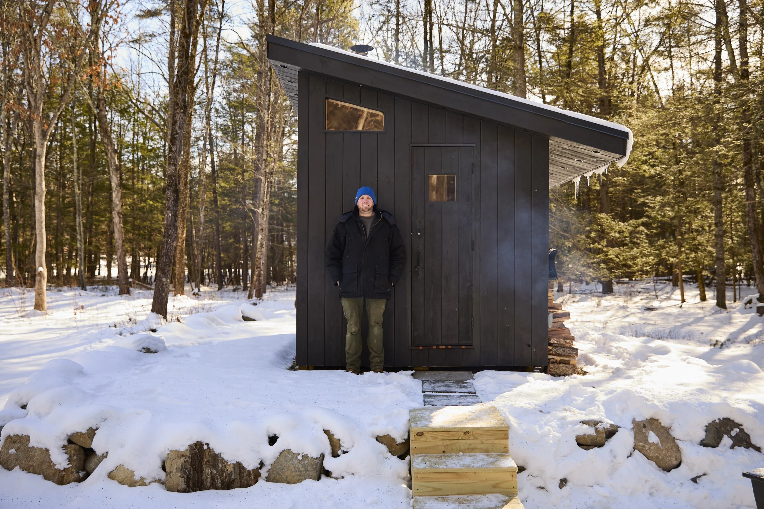 Man in a blue beanie standing outside a modern black tiny house in a snowy forest