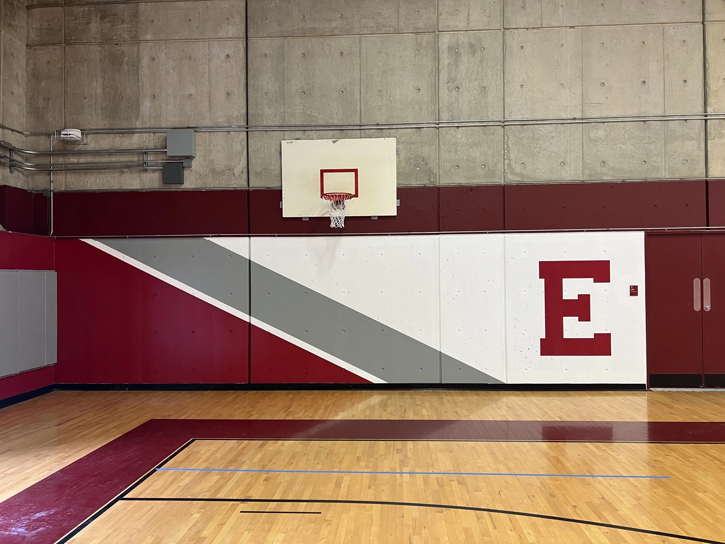 Indoor basketball court with a hoop, polished hardwood floor, and maroon-and-white wall featuring a large letter E.