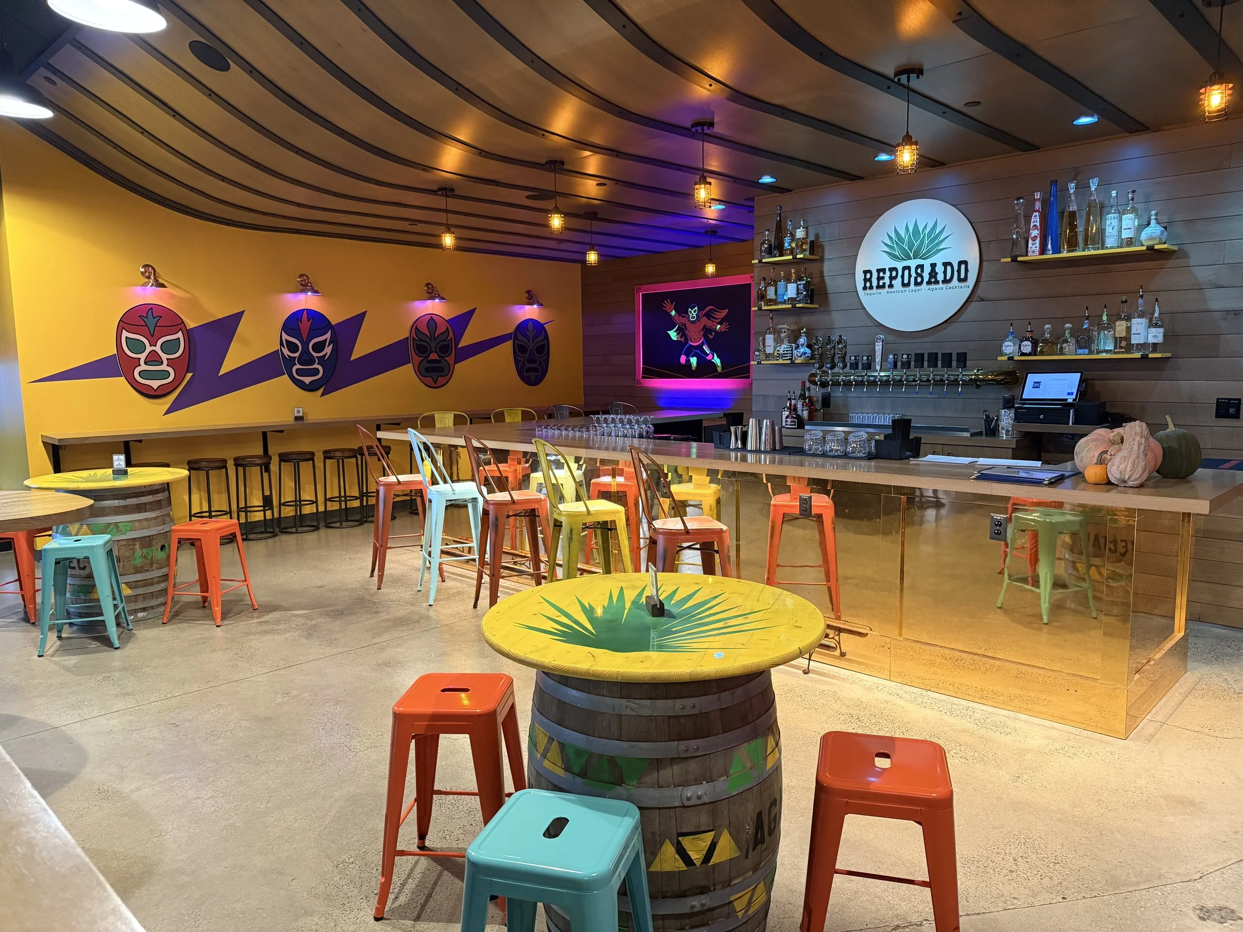 Colorful Reposado tequila bar with liquor shelves, draft taps, Mexican wrestling mask murals, and barrel tables with stools.