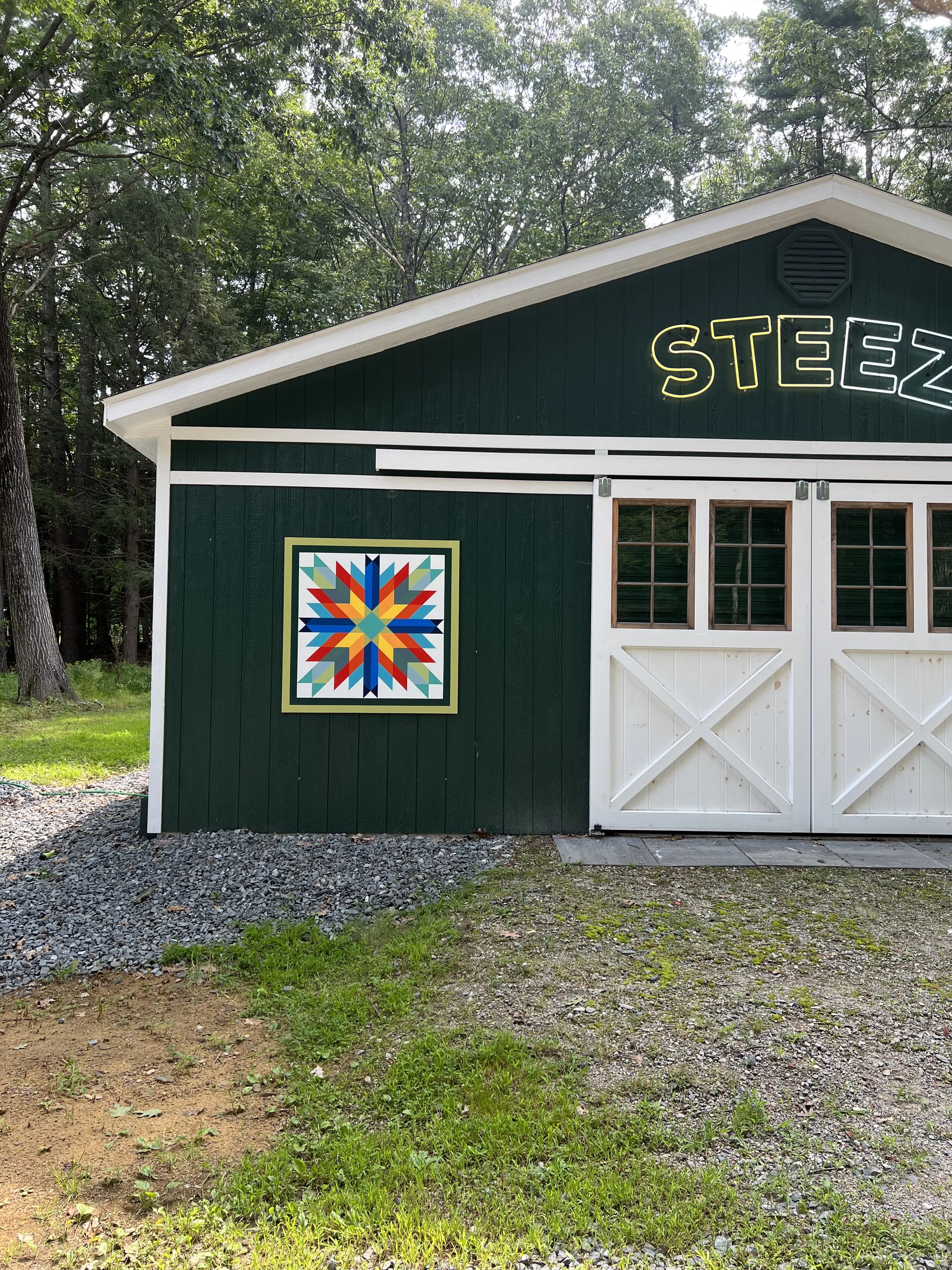 Dark green garden shed with white trim, colorful quilt-pattern wall decor, and partial neon-style lettering surrounded by trees.