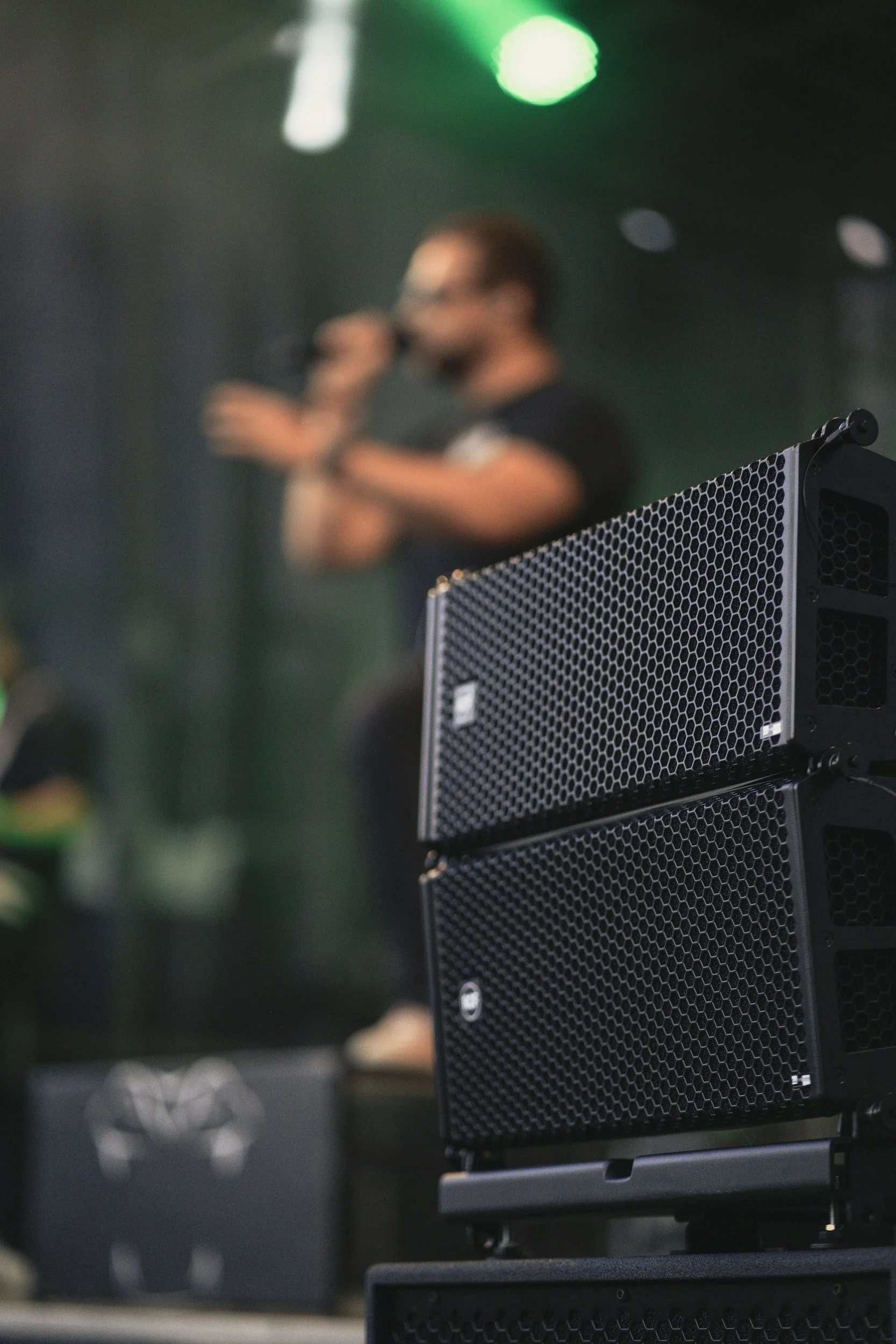 A blurred person with glasses is speaking into a microphone in the background. In the foreground, a speaker or amplifier with a black metal grille is prominently in focus.
