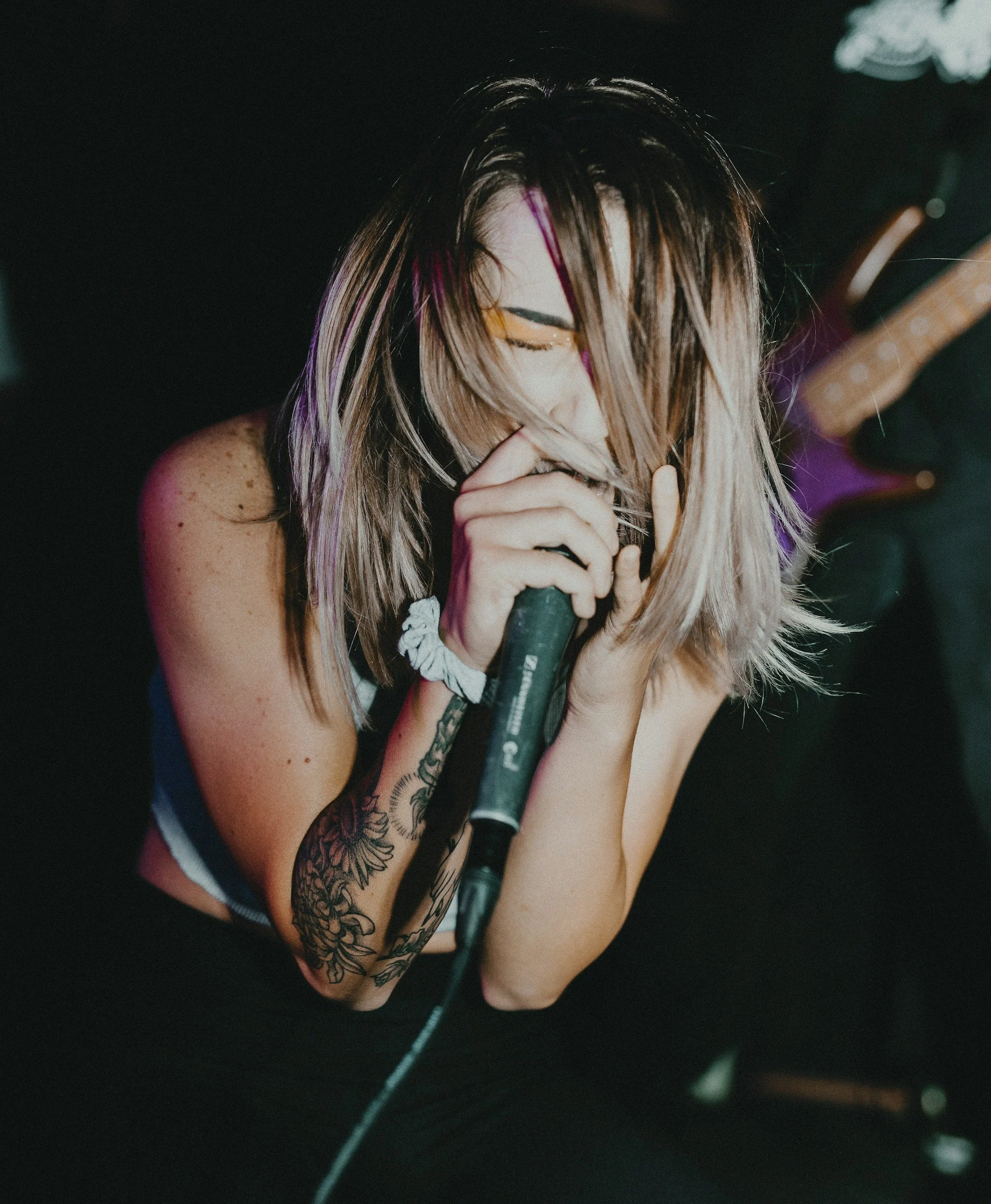 A woman with long blonde hair, tattoos on her arm, and wearing a black tank top with colorful stripes, holding a microphone and singing with her eyes closed.