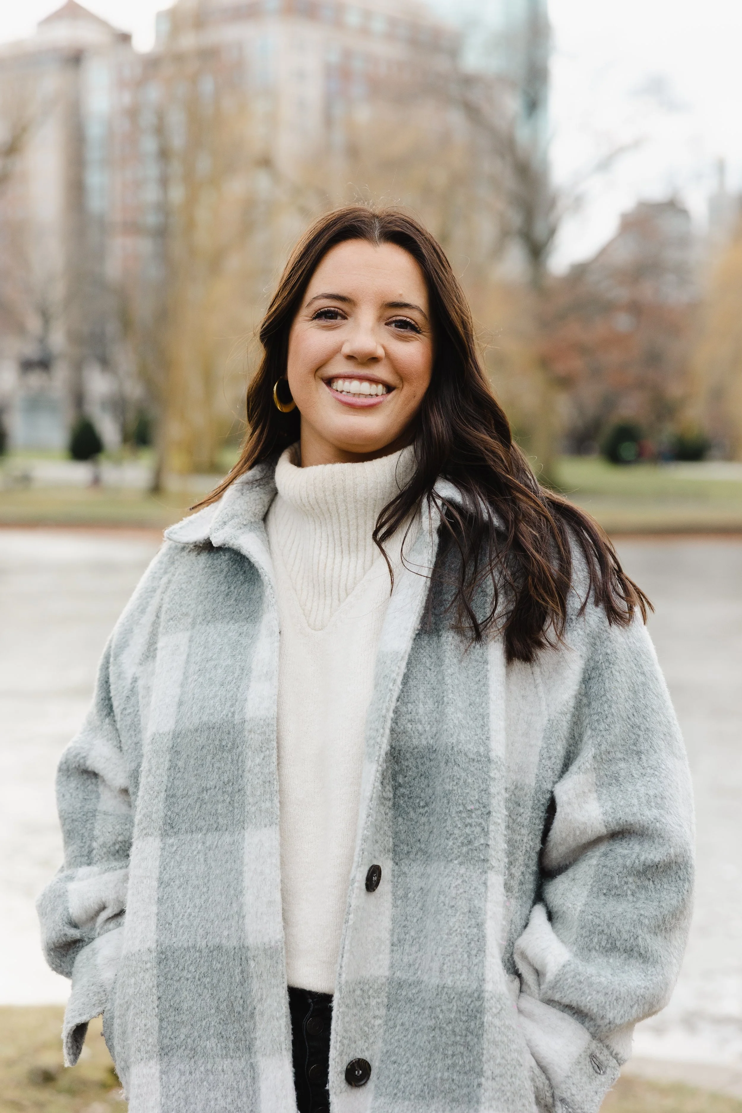 A woman with long brown hair smiling outdoors in autumn, wearing a light gray plaid coat and a white turtleneck sweater, with trees and buildings in the background.