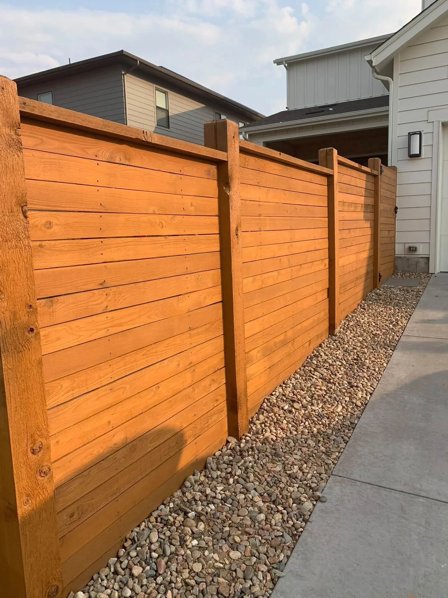 A wooden privacy fence next to a concrete sidewalk and a gravel strip, with suburban houses in the background under a partly cloudy sky.