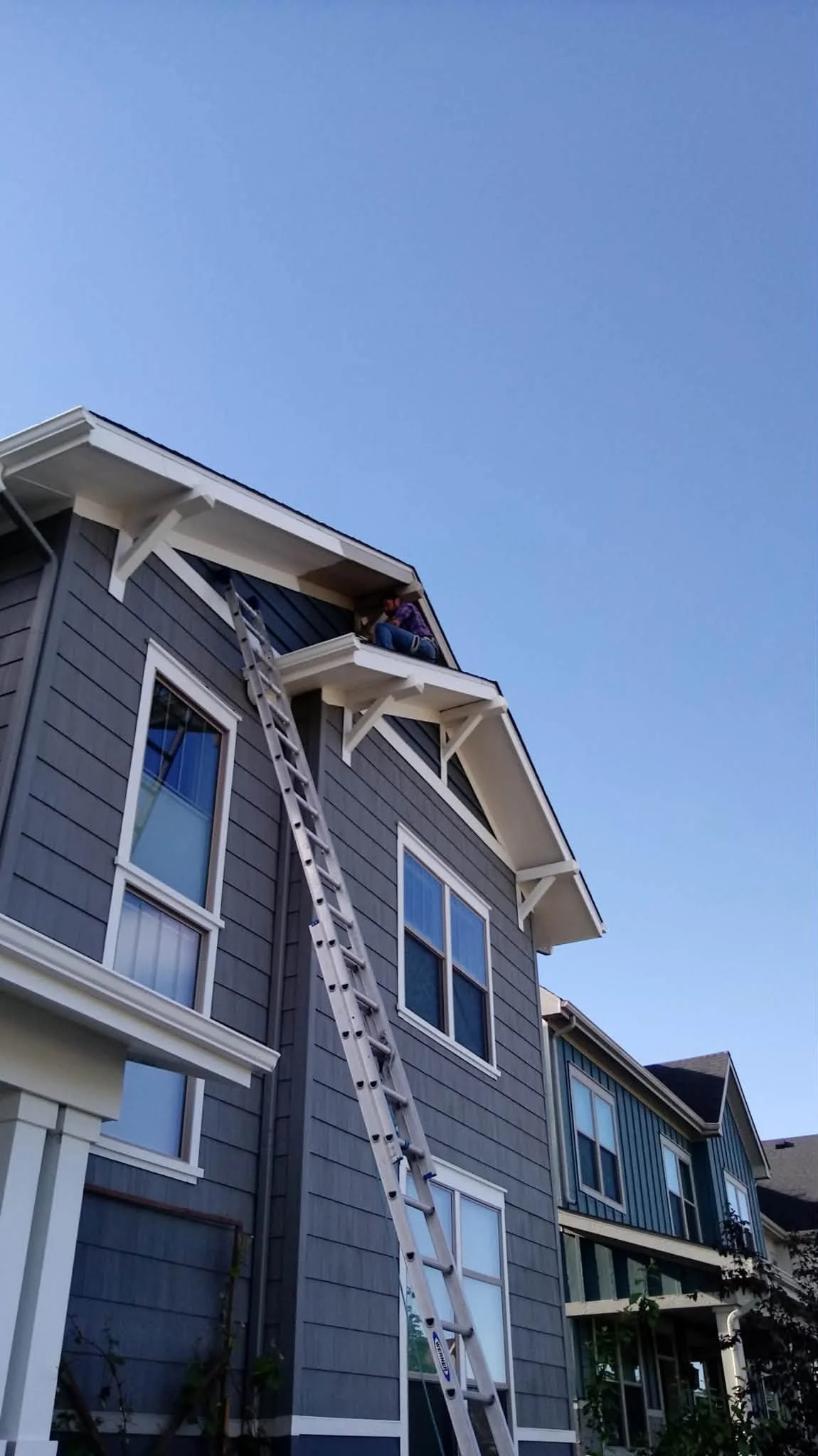 A person sitting on a balcony of a modern grey apartment building, reaching towards the roof with a ladder leaning against the building, under a clear blue sky.