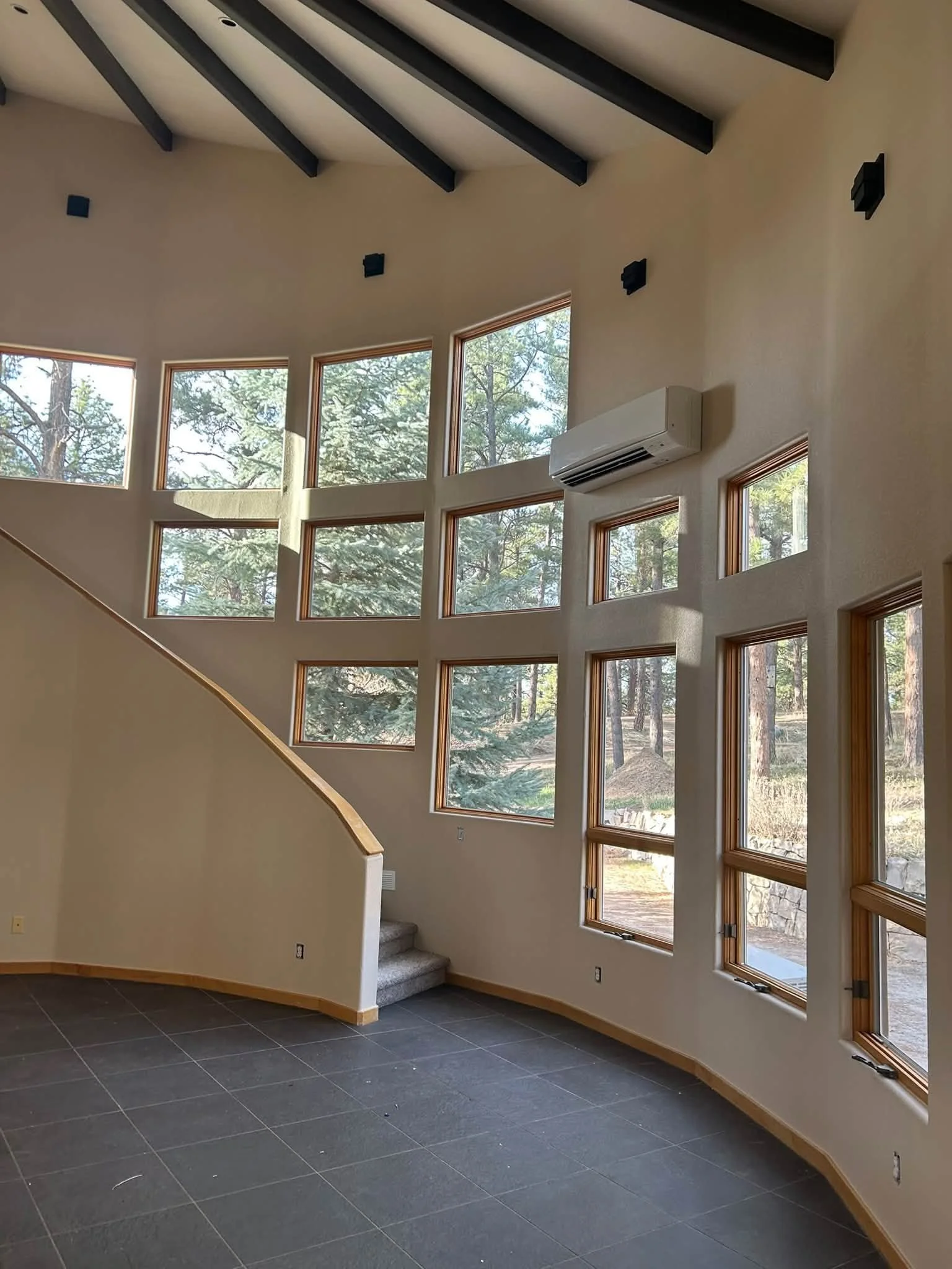 Interior view of a room with a curved wall featuring numerous windows showing trees outside, a staircase with carpeted stairs, and an air conditioning unit mounted on the wall.