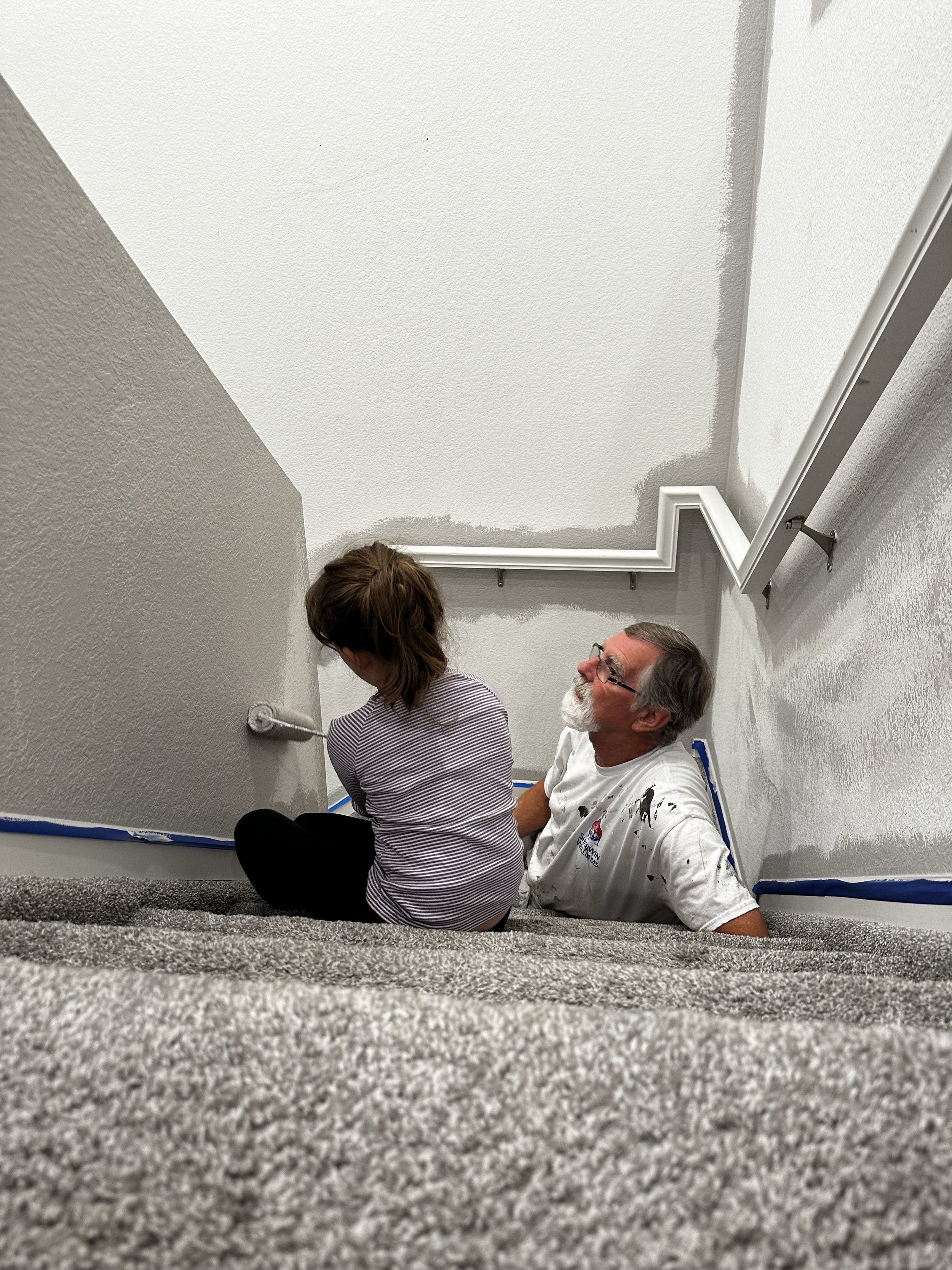 Two people sit on a carpeted floor against a wall, with painters tape along the baseboard. One person has brown hair and is wearing a striped shirt, while the other has gray hair, glasses, and a white paint-stained shirt. They appear to be taking a break during painting.