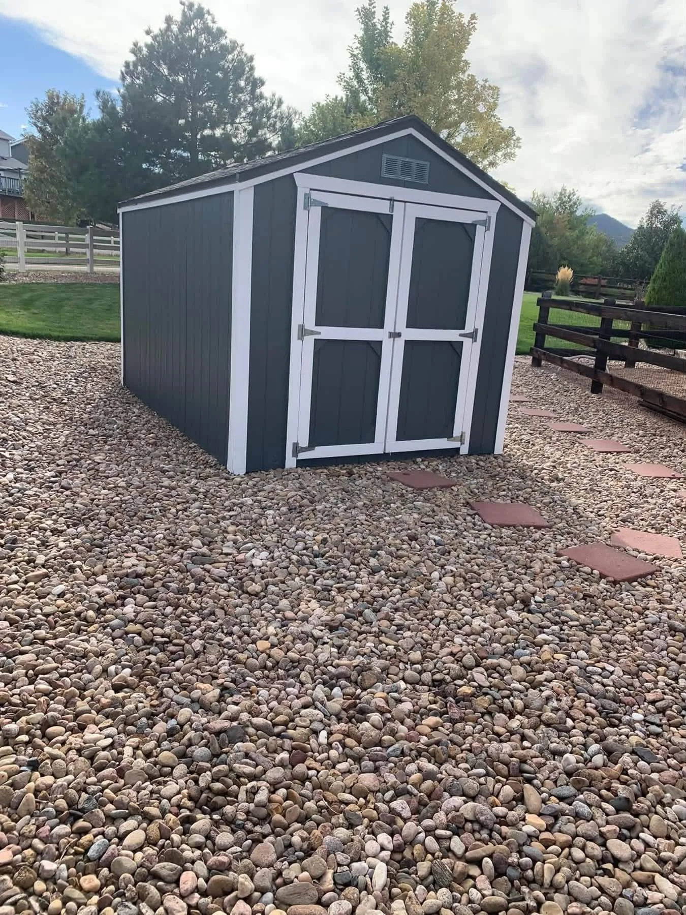 A small black garden shed with white trim on a bed of multicolored pebbles, with a pathway of stepping stones nearby, greenery, and trees in the background under a partly cloudy sky.