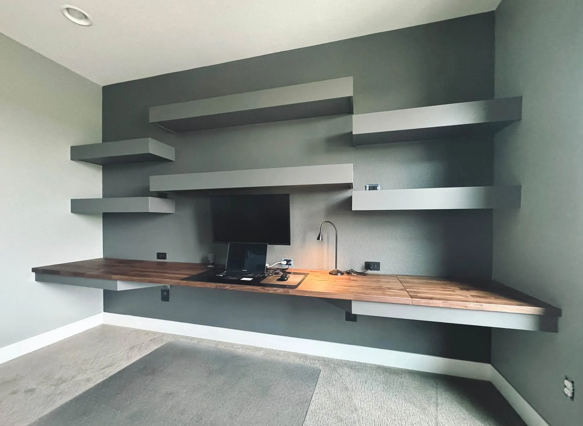 A minimalist room with floating gray shelves on the wall, a wooden desk with a computer monitor, a lamp, and a carpeted floor.