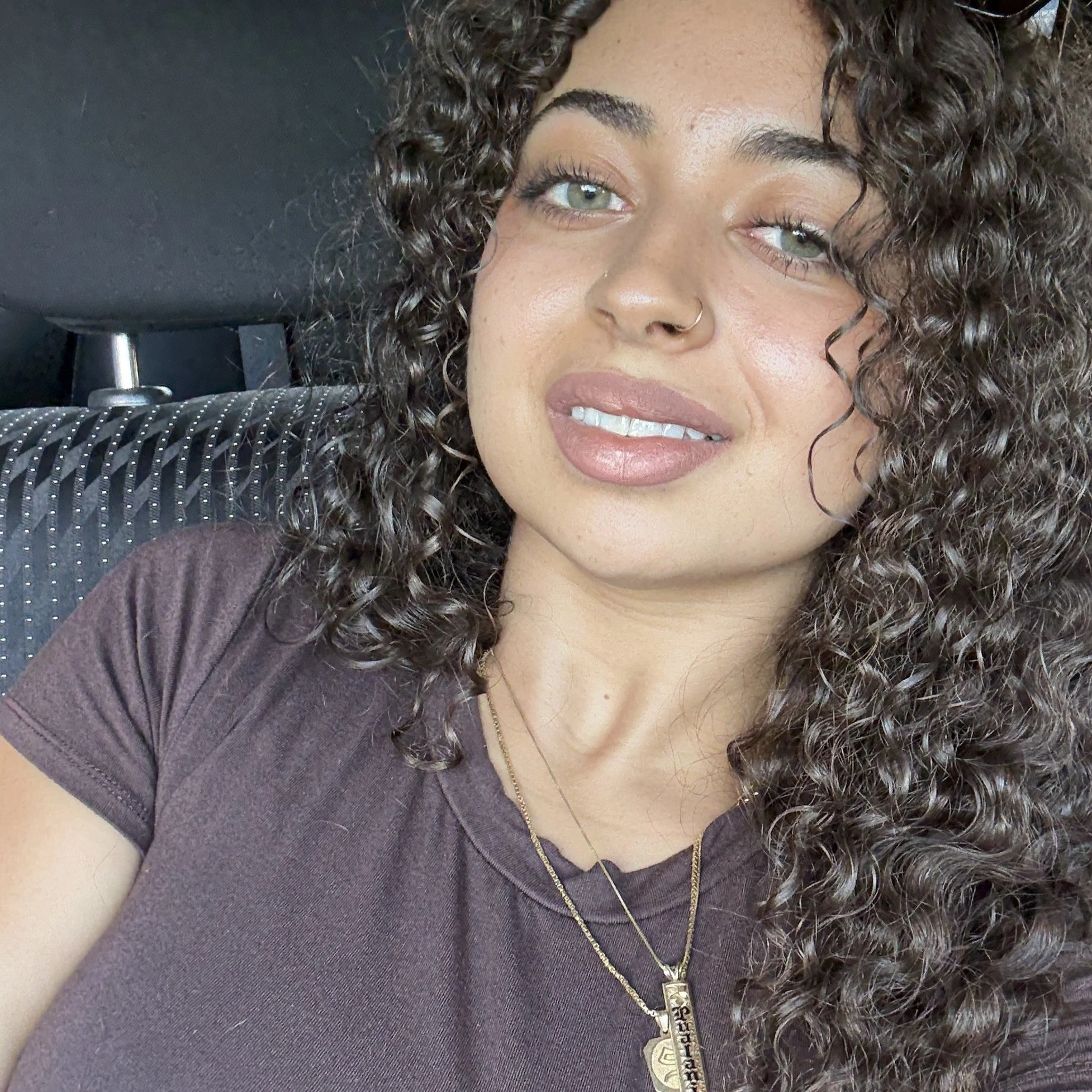 Close-up photo of a woman with curly hair, light skin, wearing a dark top with pinstripes, gold layered necklaces, and a nose piercing.