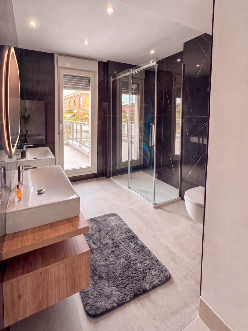 Baño moderno con ducha de cristal, lavabo blanca sobre mueble de madera, espejo con luz, ventana que da al exterior, piso de baldosas claras y alfombra gris