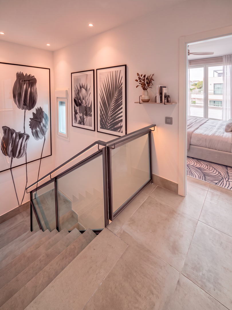 Imagen de un espacio interior de una vivienda que incluye una escalera de vidrio y metal, paredes blancas decoradas con cuadros en tonos negros y un pasillo que conduce a una habitación con cama y ventanales que permiten la entrada de luz natural.