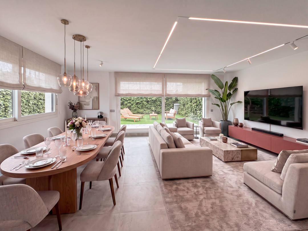 Interior moderno de sala y comedor con grandes ventanales, muebles beige y un espacio verde exterior visible por la ventana.