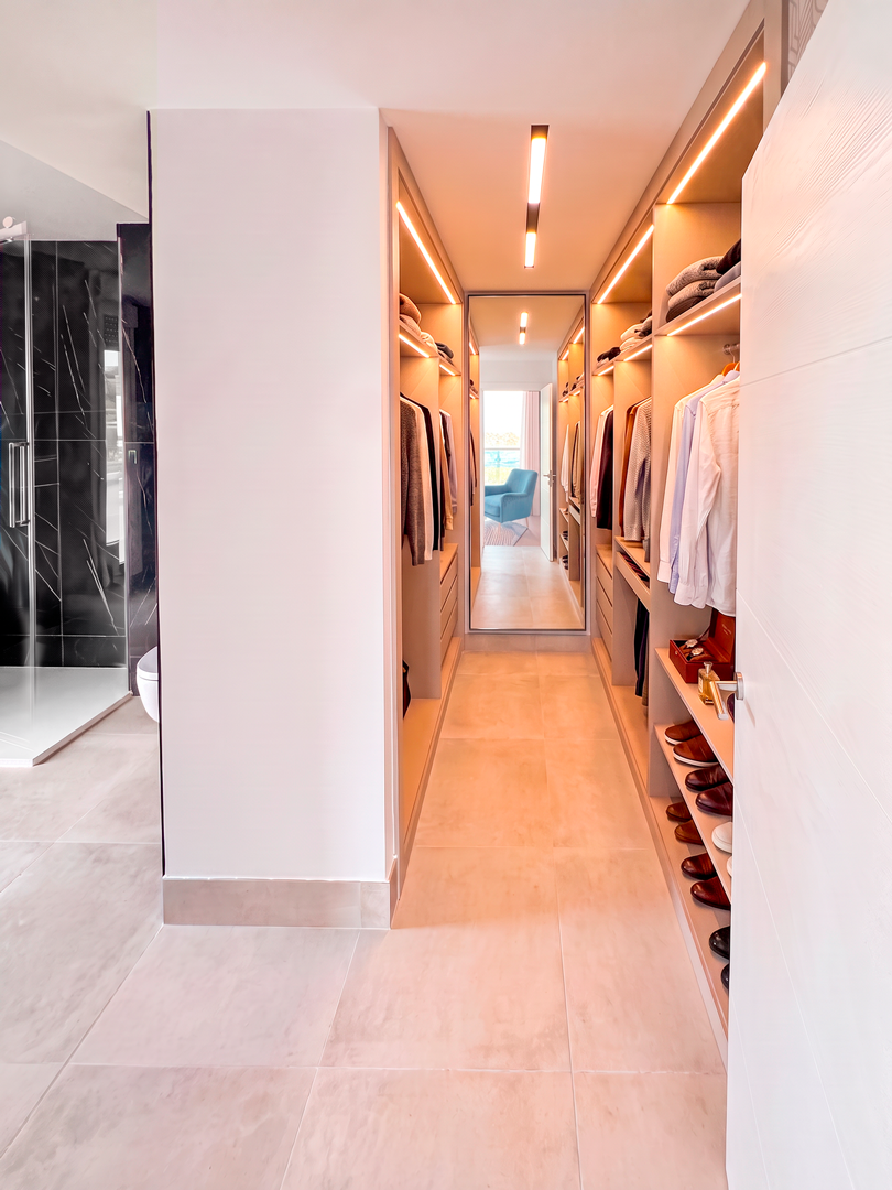 Vestidor con estanterías llenas de ropa y zapatos, con un espejo grande en el fondo y luz cálida en el techo.