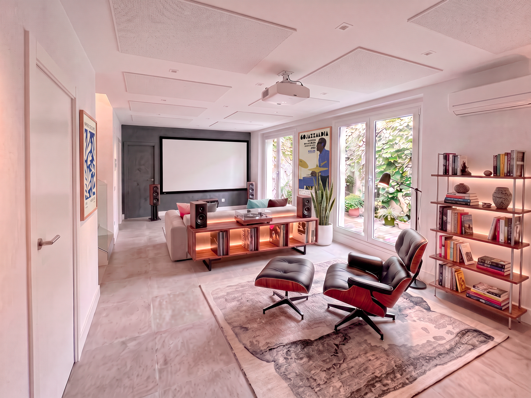 Sala de estar moderna con sillones de cuero negro, mesa de madera, estantería con libros, plantas, pantalla de proyector y ventanas con vista a un jardín.