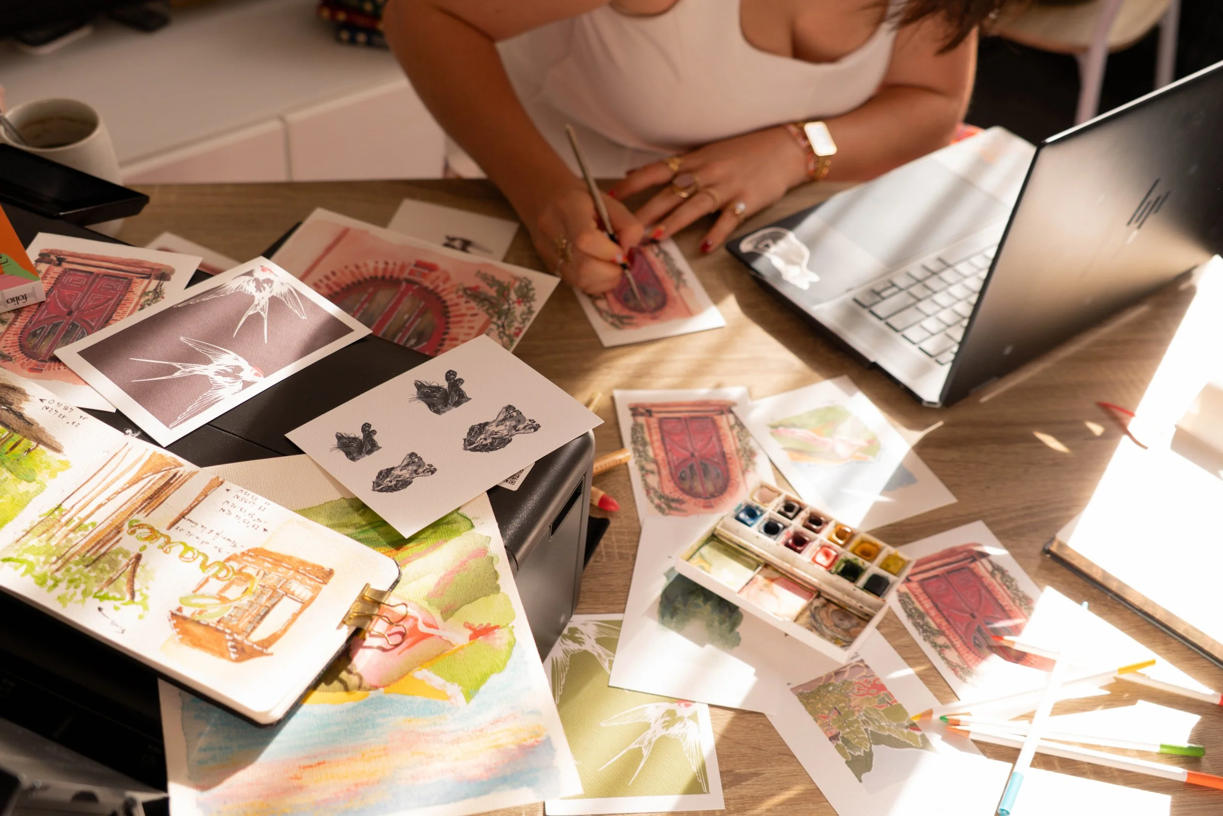 Une personne travaillant sur de nombreux dessins et aquarelles colorés sur un bureau en bois, avec un ordinateur portable et des petits pots de peinture.