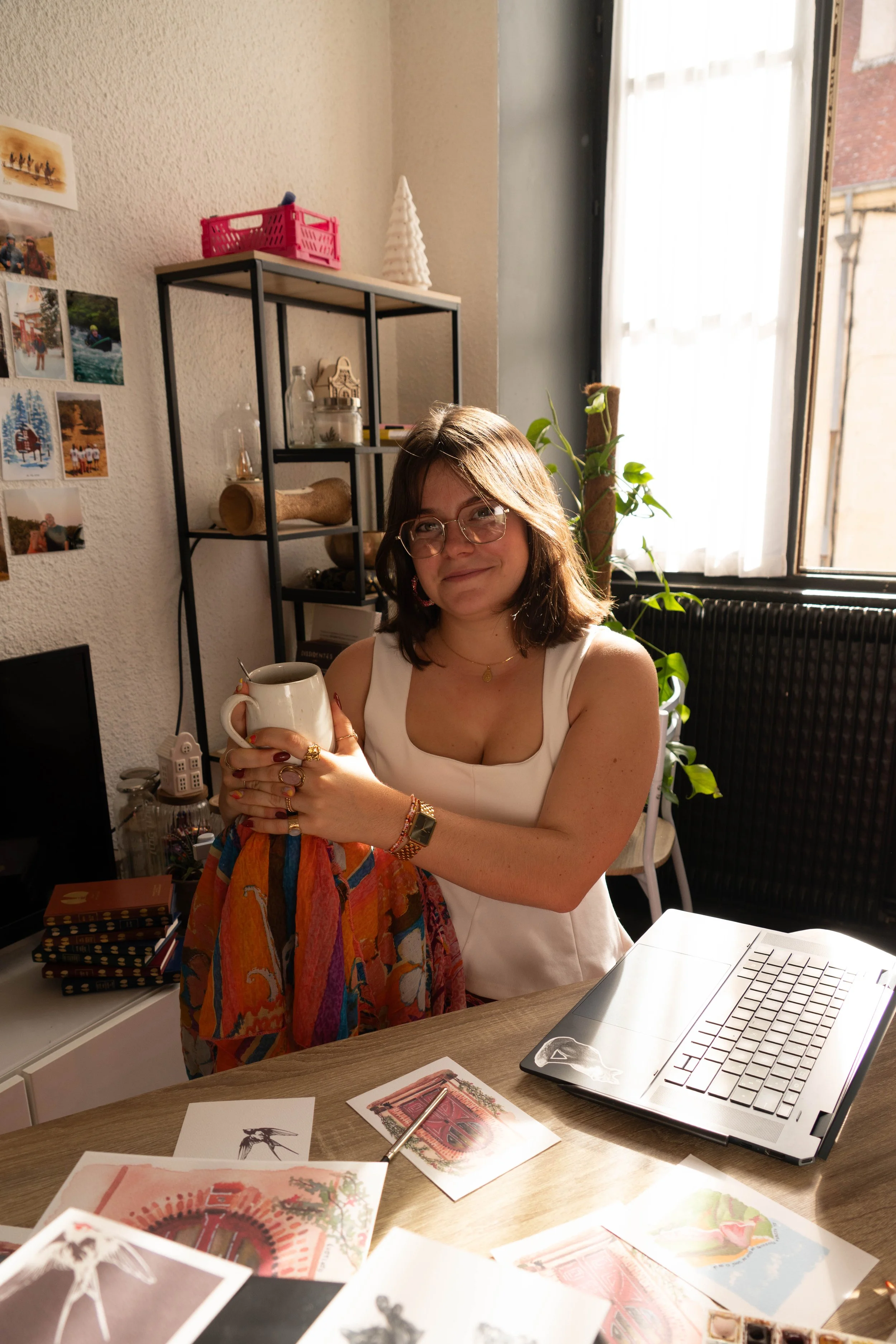 Une jeune femme souriante tient une tasse dans un bureau lumineux, entourée de croquis artistiques, d’un ordinateur portable et de décorations comme une guirlande lumineuse et une plante d'intérieur.