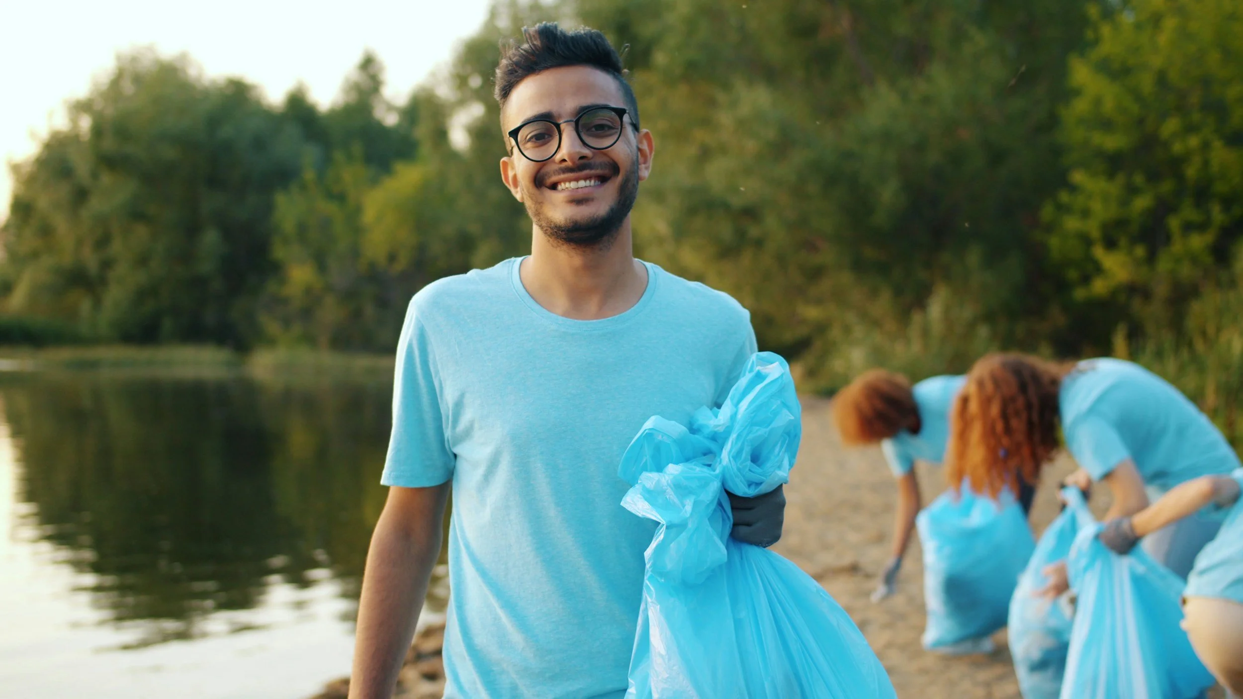 A young man smiling at the camera while holding a large blue trash bag during a cleanup event by a river, with other volunteers in the background collecting trash.
