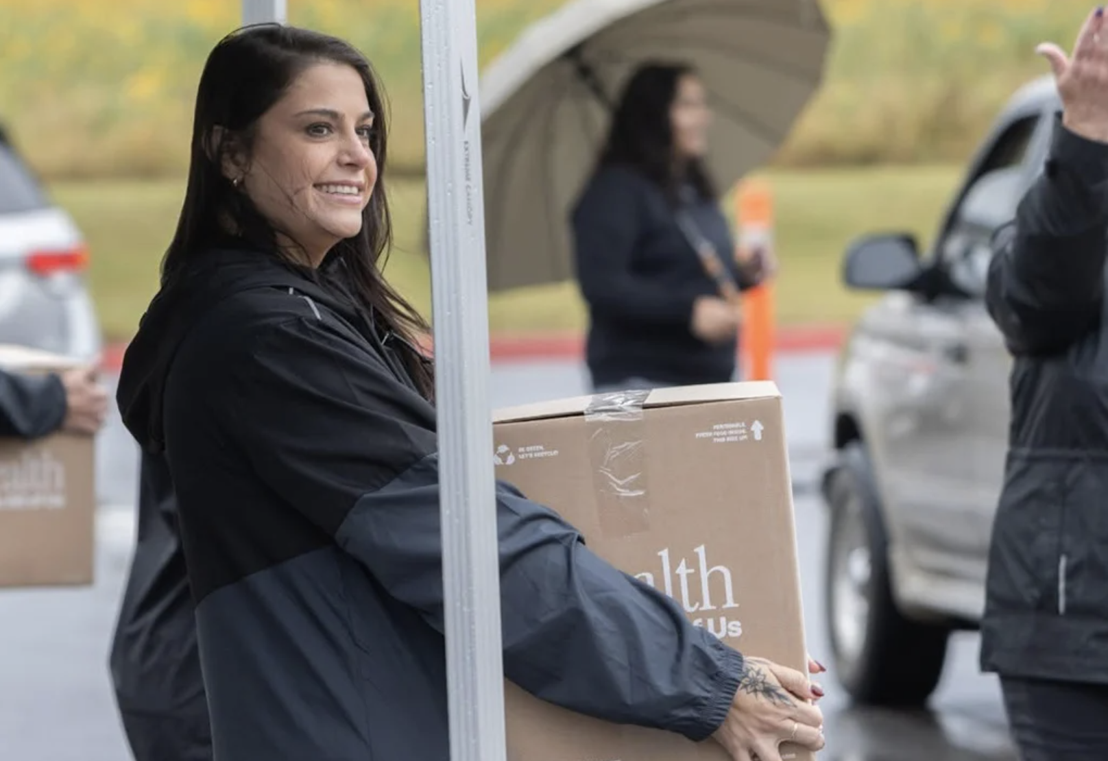 A woman with long dark hair and tattoos on her hand holding a large box at a donation or distribution event outdoors in rainy weather, with other people and cars visible in the background.
