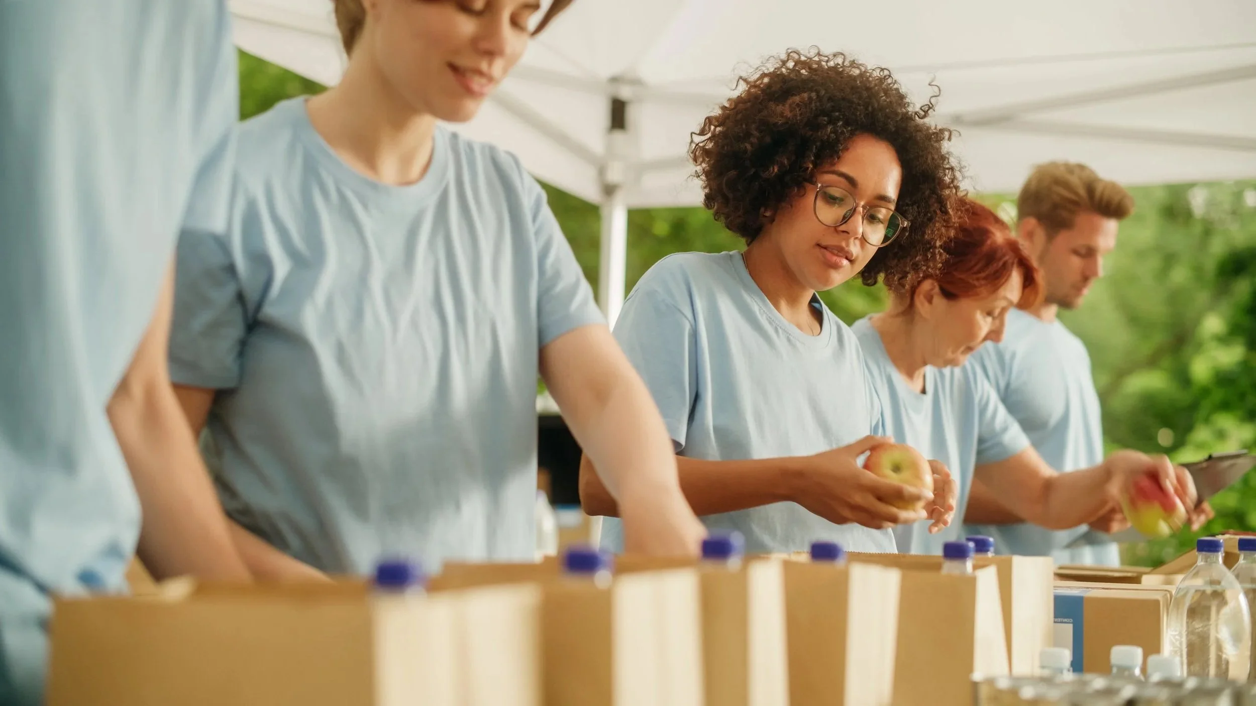 Group of people in light blue shirts working with apples at an outdoor event under a white canopy.