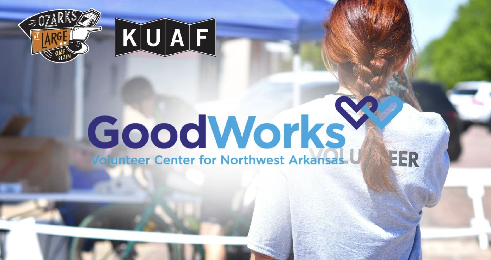 Back view of a volunteer with a red braid, wearing a gray shirt, at the GoodWorks Volunteer Center for Northwest Arkansas, with logos of Ozarks at Large and KUAF in the background.