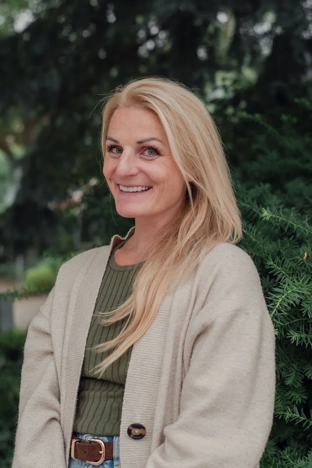 A woman with long blonde hair smiling outdoors, wearing a beige cardigan over a green top, with greenery in the background.