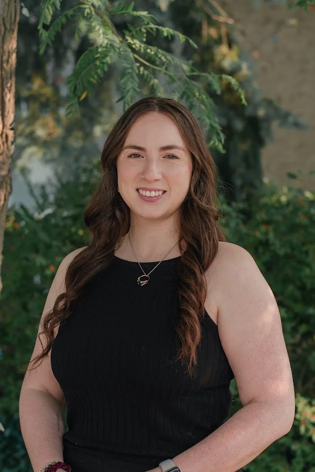 Young woman with long brown hair smiling outdoors, standing in front of green plants and trees.