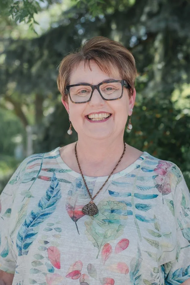 A smiling woman with short brown hair, wearing large black glasses, earrings, a necklace, and a white shirt with colorful leaves printed on it, outdoors with green trees in the background.