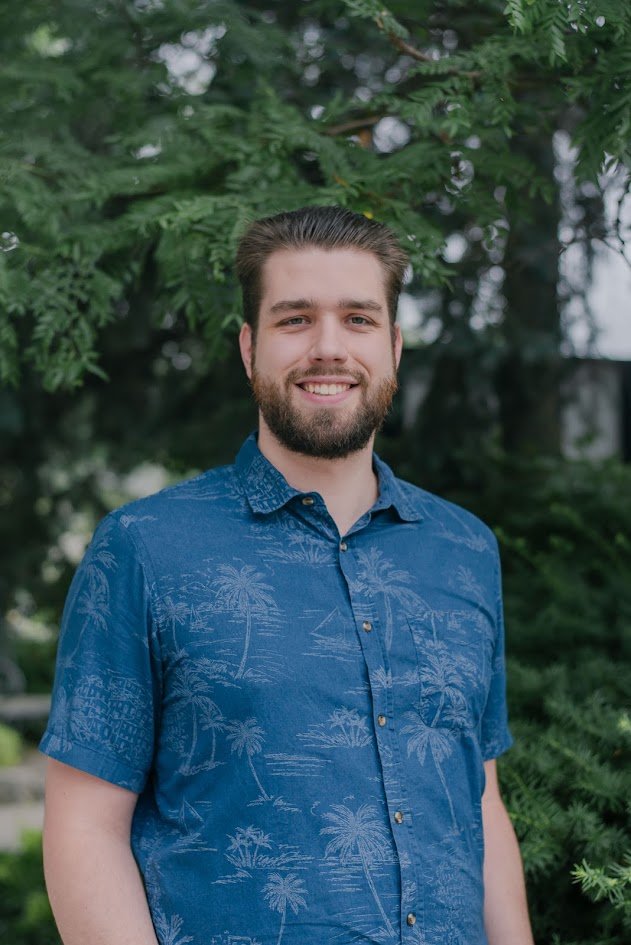 A young man with a beard and brown hair smiling outdoors, wearing a blue button-up shirt with a palm tree pattern, standing in front of green trees.