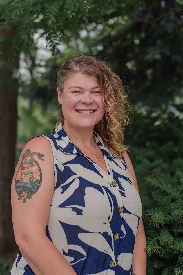 A smiling woman with curly hair and a tattoo on her upper arm, standing outdoors with green foliage in the background.