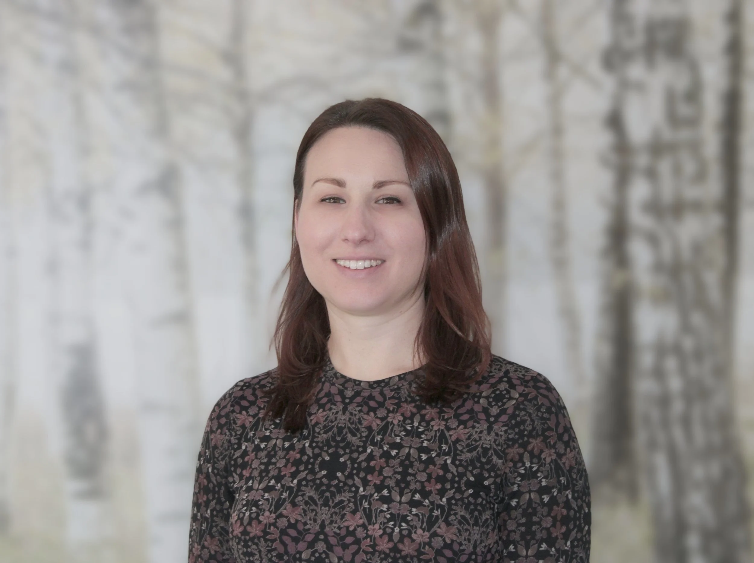 Portrait of a woman with shoulder-length brown hair and a smile, wearing a black dress with a floral pattern, standing against a blurred background of trees.
