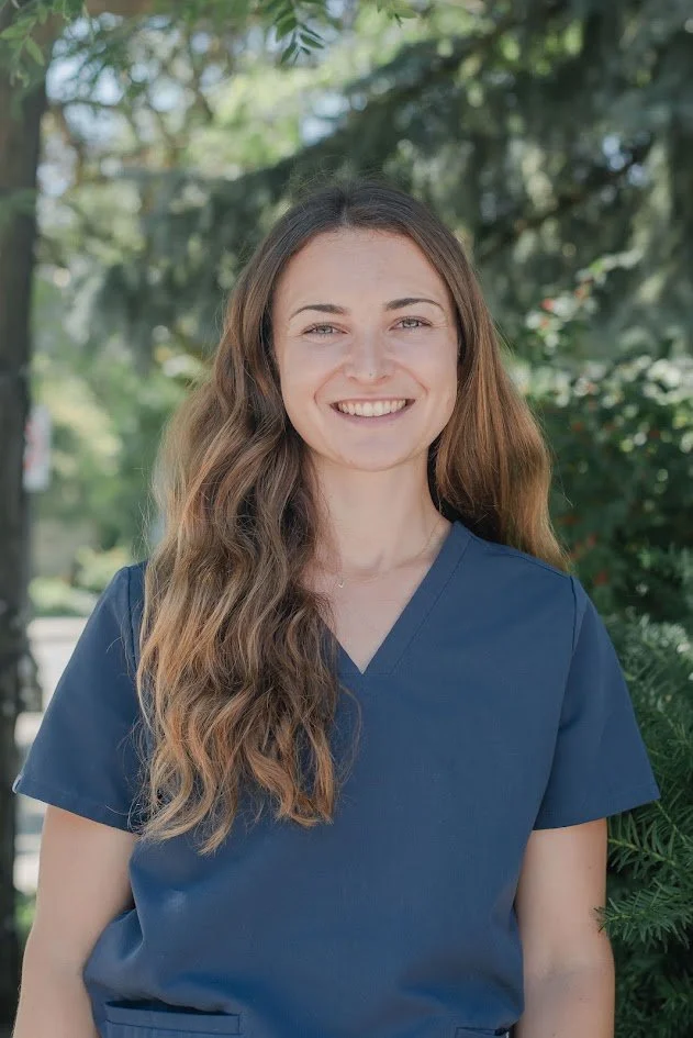 A young woman with long, wavy brown hair is smiling outdoors, wearing navy blue scrubs, with green trees in the background.