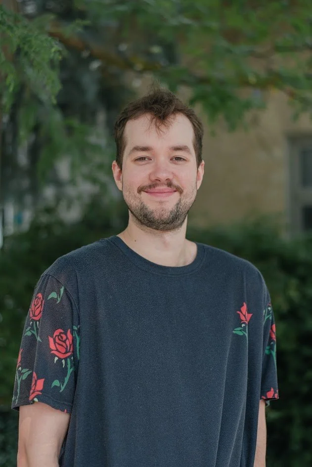 A young man with brown hair and a beard smiling outdoors, wearing a dark blue T-shirt with red roses on the sleeves, with trees and a building in the background.