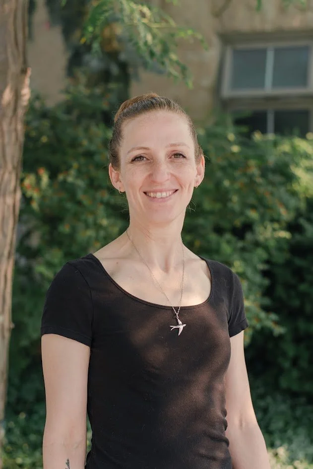 A woman with light skin and brown hair tied back, smiling outdoors, wearing a black t-shirt and a bird-shaped pendant necklace, with greenery and a building in the background.