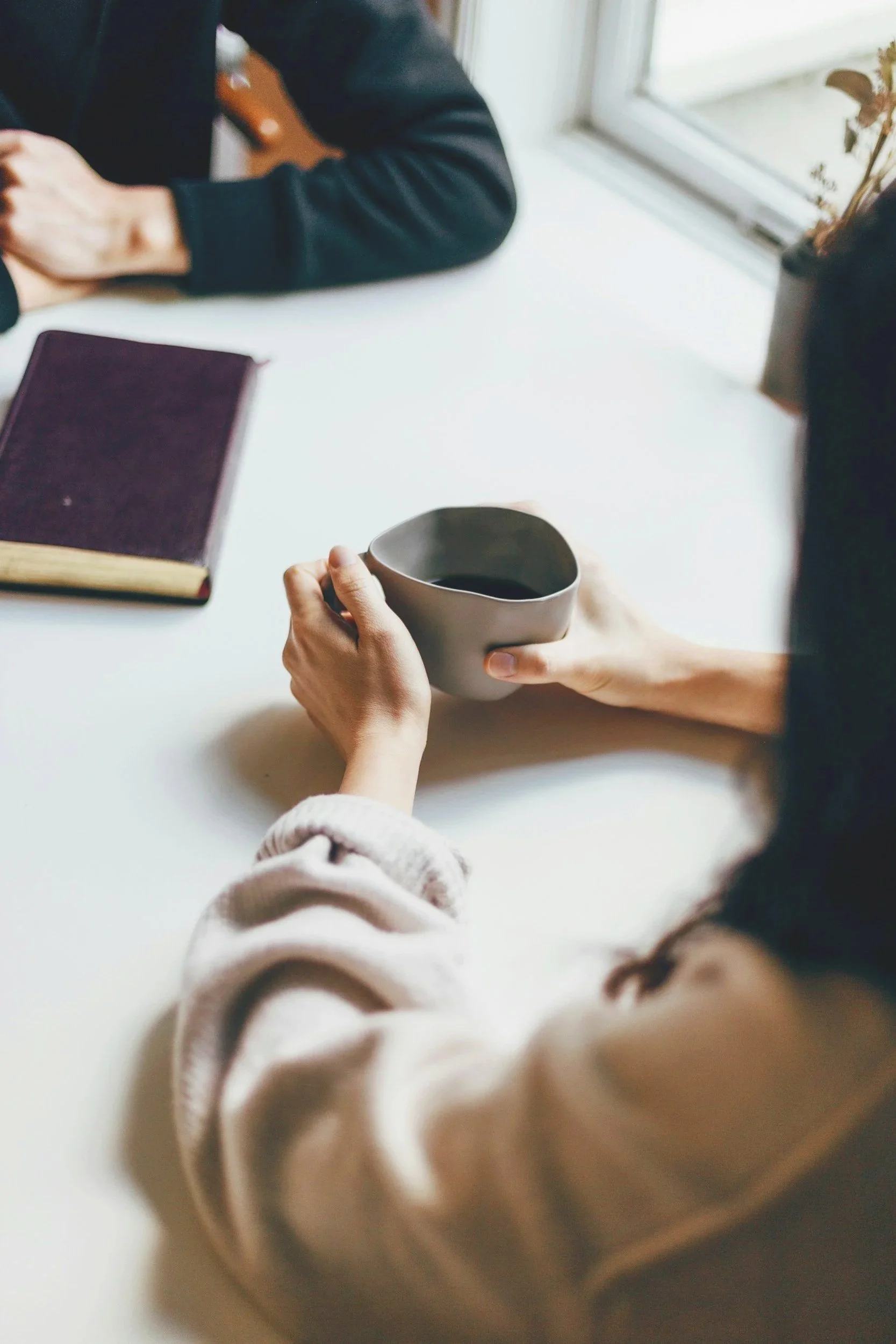 Close-up of a person's hands holding a gray mug filled with dark coffee, with a blurred background including a person wearing a dark jacket and a notebook on a white table near a window.
