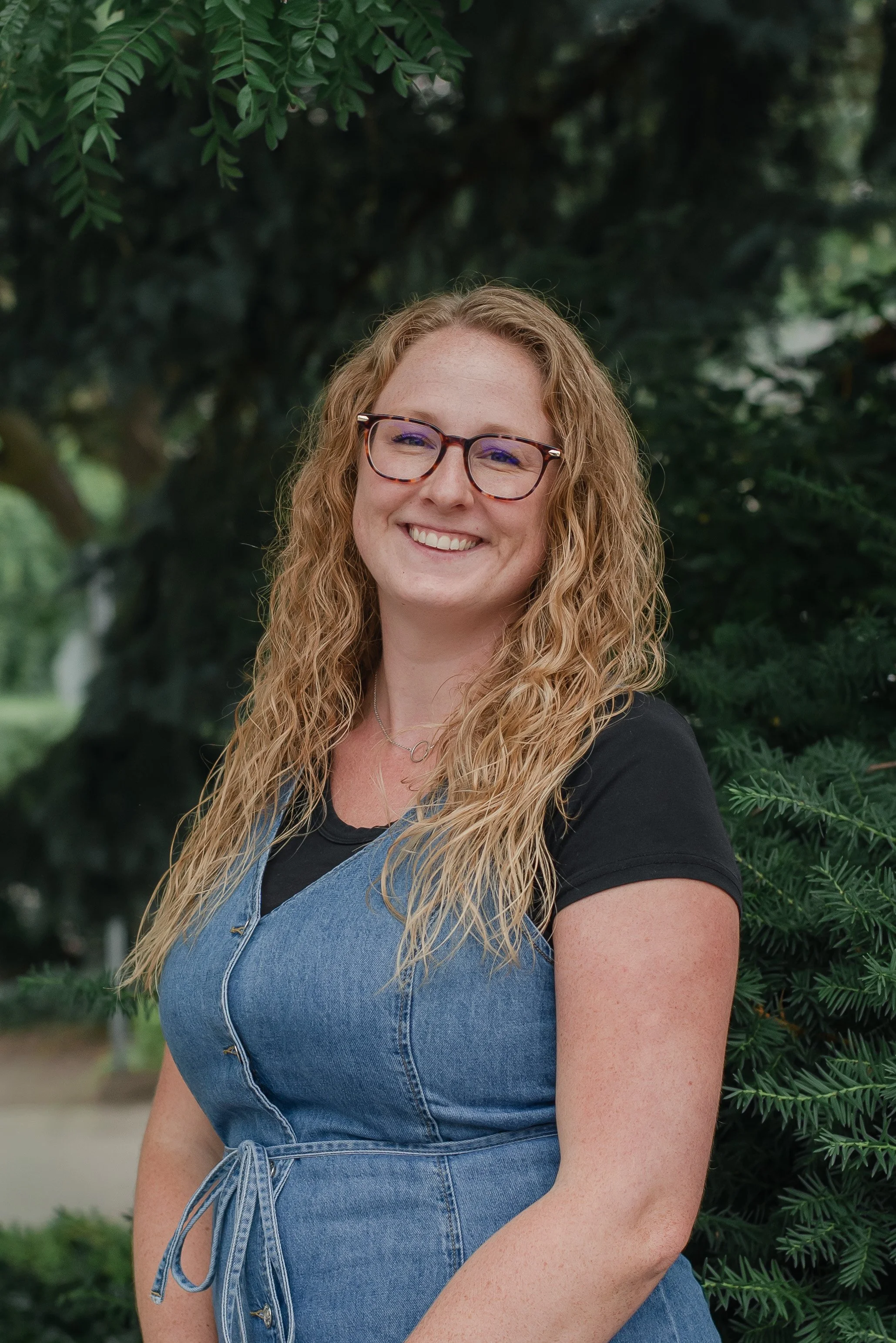 A young woman with long curly blonde hair, glasses, and a black t-shirt, smiling outdoors with greenery in the background.