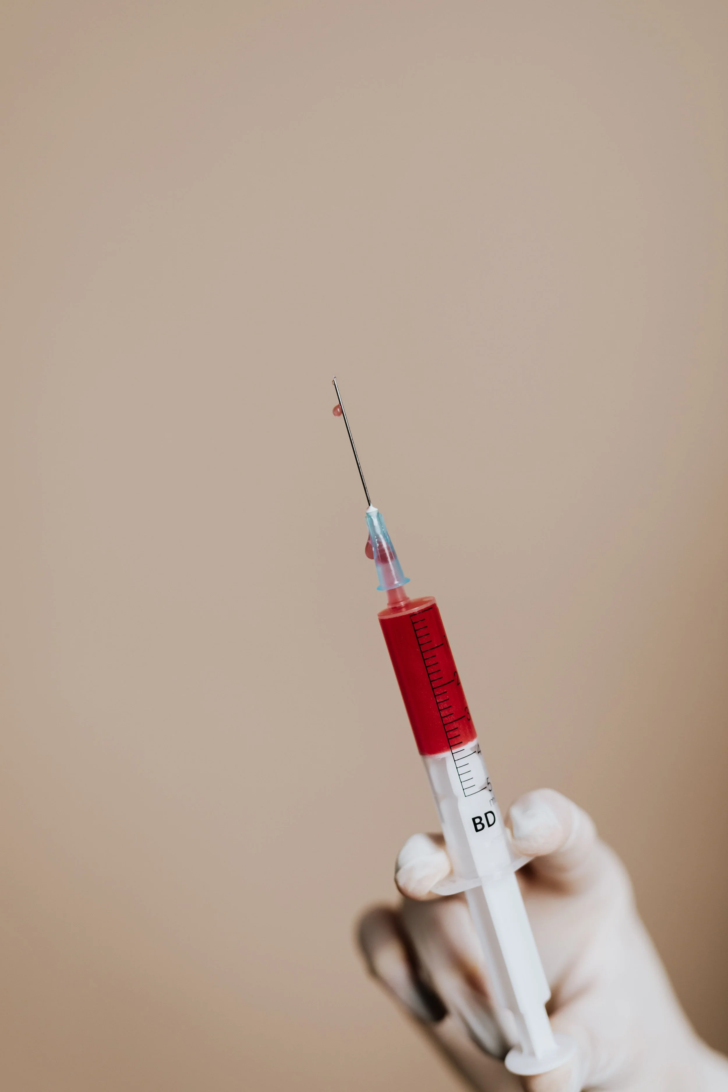 A healthcare professional's hand wearing a glove holding a syringe filled with red liquid, with a beige background.