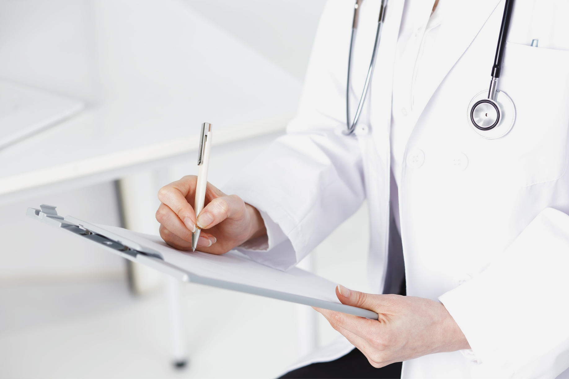 A healthcare professional in a white coat with a stethoscope around their neck writing on a clipboard with a pen.