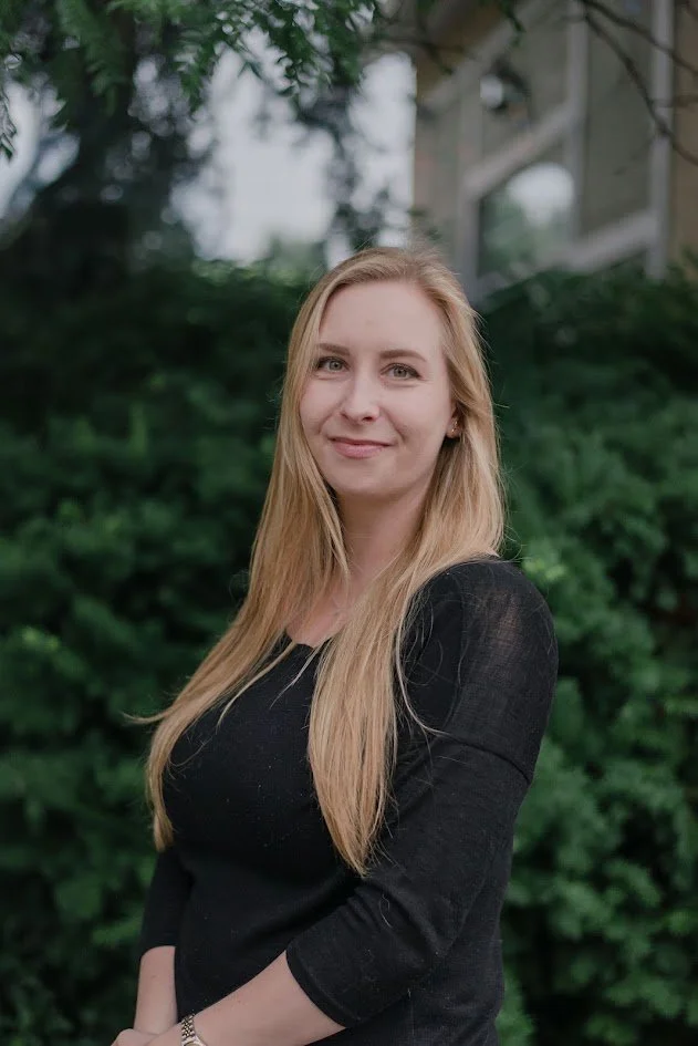 A young woman with long blonde hair, wearing a black top, standing outdoors with greenery in the background and a building structure in the distance, smiling softly at the camera.