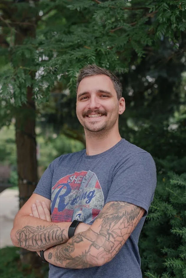 A young man with short brown hair, beard, and tattoos on his arms, smiling with arms crossed in front of a green leafy background.
